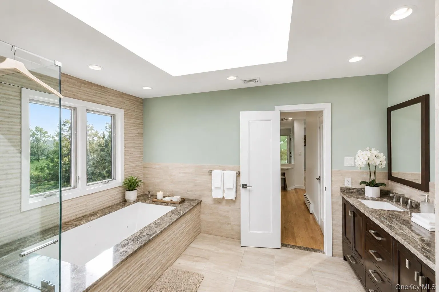Bathroom featuring a built-in soaking tub with stone surround, a separate glass-enclosed shower, a double window, and a dark wood vanity with a stone countertop and dual sinks Bathroom featuring a built-in soaking tub with stone surround, a separate glass-enclosed shower, a double window, and a dark wood vanity with a stone countertop and dual sinks