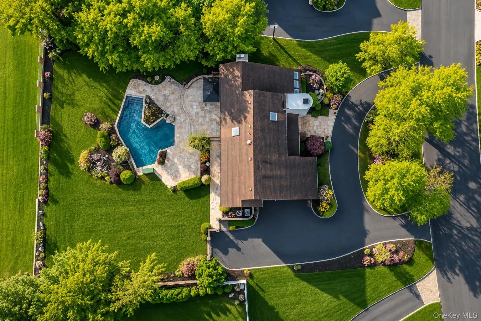Expansive property featuring a dark asphalt driveway, a large in-ground swimming pool with surrounding stone patio, and mature trees and landscaping Expansive property featuring a dark asphalt driveway, a large in-ground swimming pool with surrounding stone patio, and mature trees and landscaping