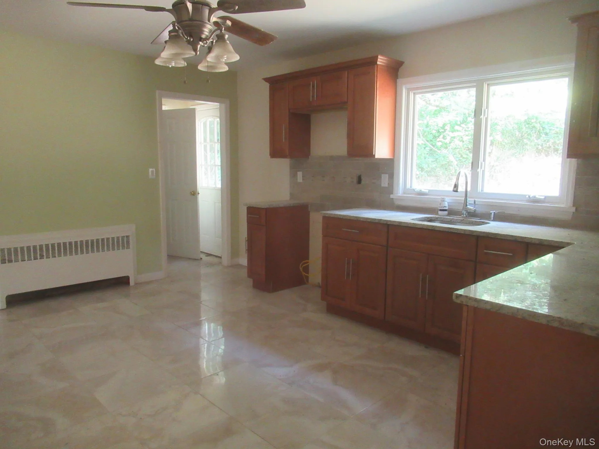 Kitchen featuring brown cabinets, radiator, tasteful backsplash, a ceiling fan, and light stone counters Kitchen featuring brown cabinets, radiator, tasteful backsplash, a ceiling fan, and light stone counters