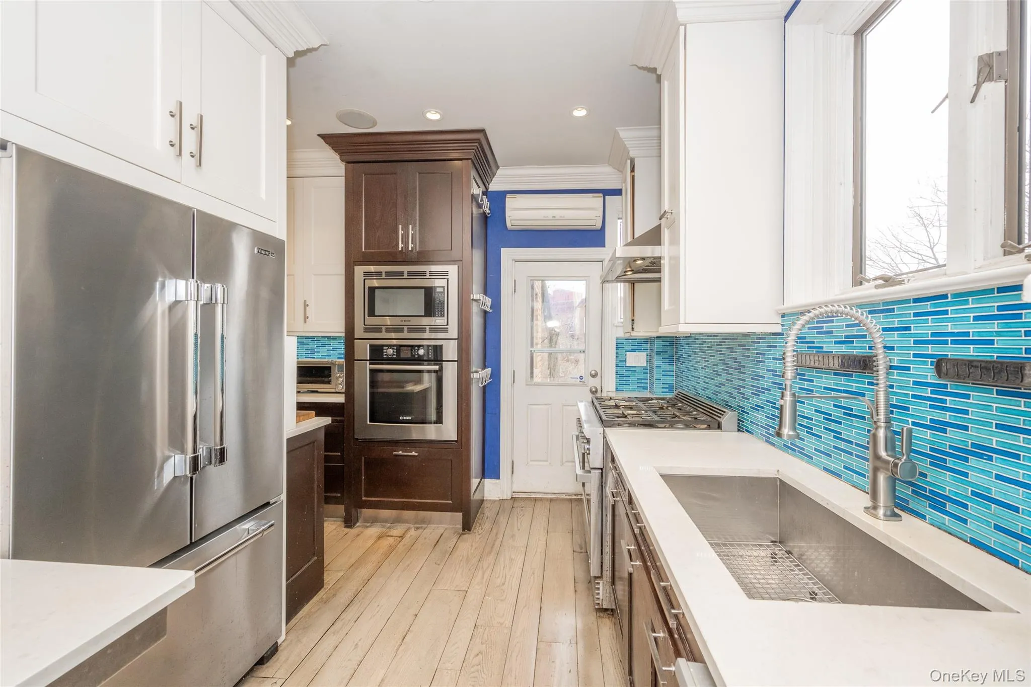 Contemporary galley kitchen featuring stainless steel appliances, wood-finish flooring, white cabinetry, integrated wall oven and microwave, and a bright blue mosaic tile backsplash Contemporary galley kitchen featuring stainless steel appliances, wood-finish flooring, white cabinetry, integrated wall oven and microwave, and a bright blue mosaic tile backsplash