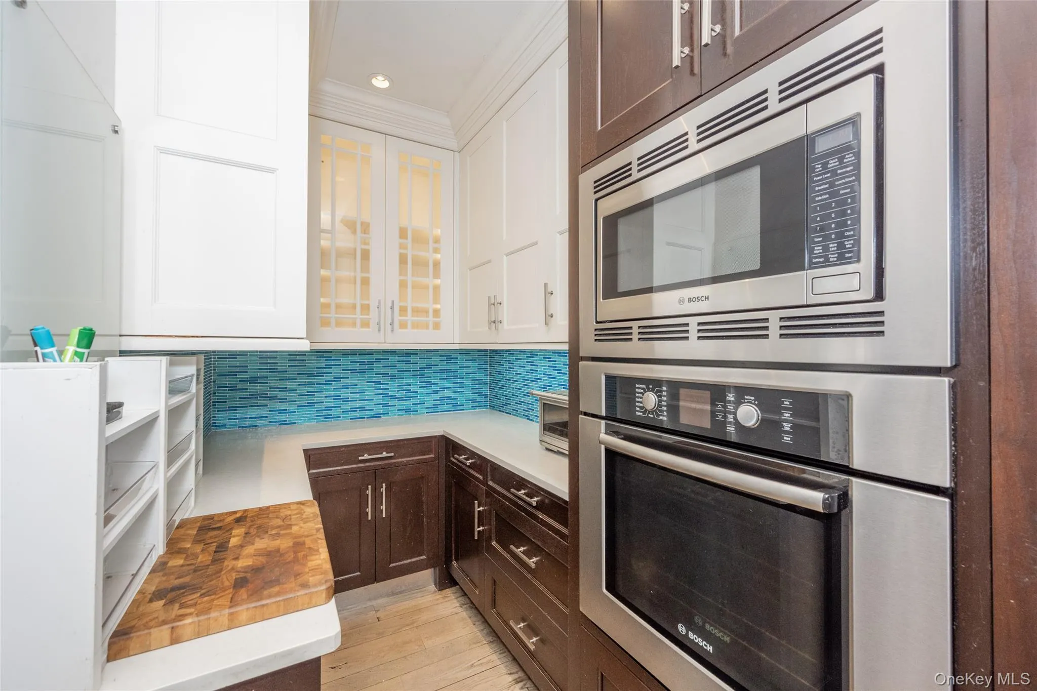 Kitchen featuring built-in stainless steel microwave and oven, two-tone cabinetry with glass-front upper cabinets, light countertop, and blue mosaic tile backsplash Kitchen featuring built-in stainless steel microwave and oven, two-tone cabinetry with glass-front upper cabinets, light countertop, and blue mosaic tile backsplash