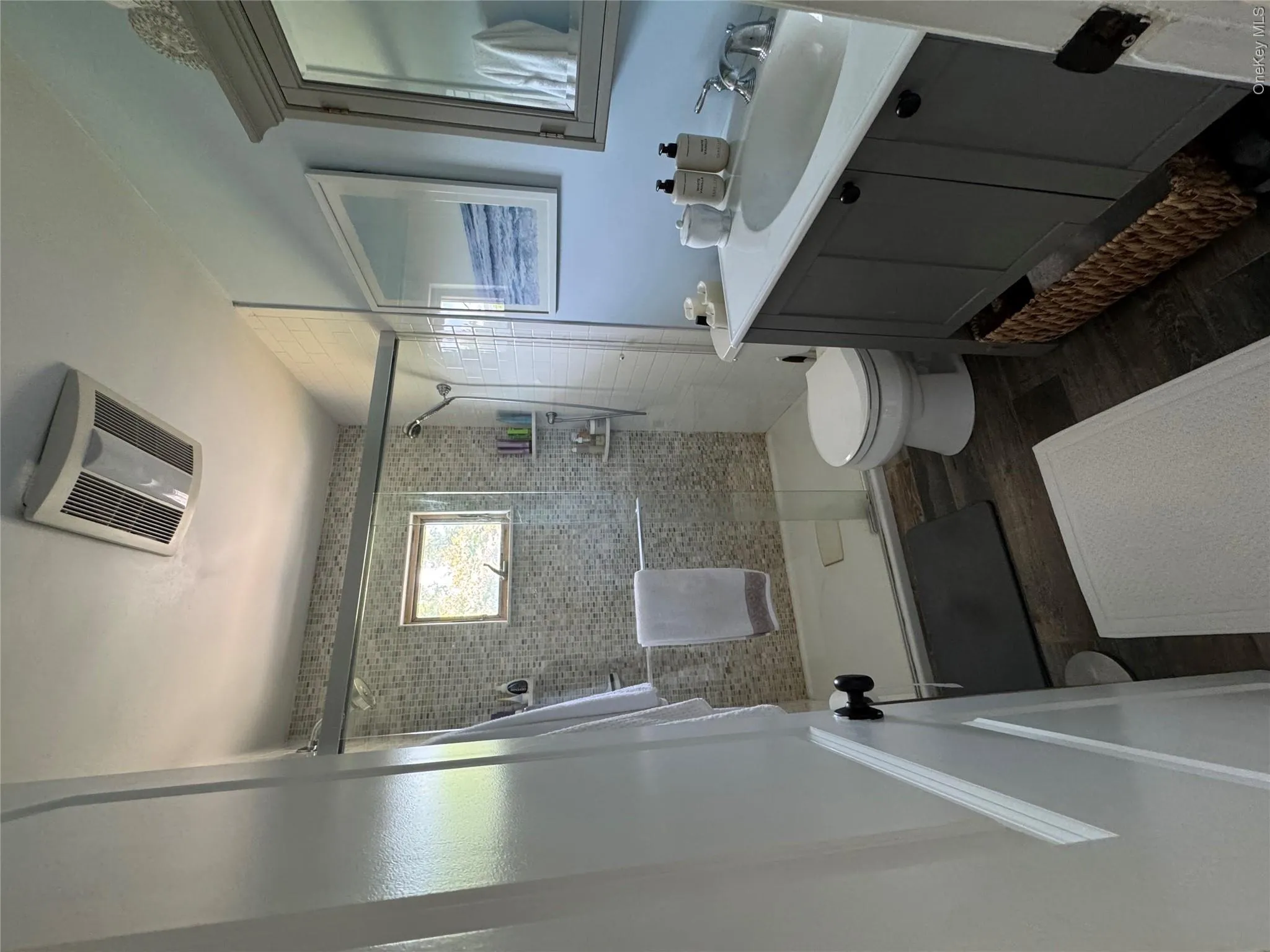 Full bathroom featuring a glass-enclosed shower with mosaic tile surround, a single vanity with a white countertop and undermount sink, wood-finish flooring, and a recessed ceiling fan with integrated lighting Full bathroom featuring a glass-enclosed shower with mosaic tile surround, a single vanity with a white countertop and undermount sink, wood-finish flooring, and a recessed ceiling fan with integrated lighting
