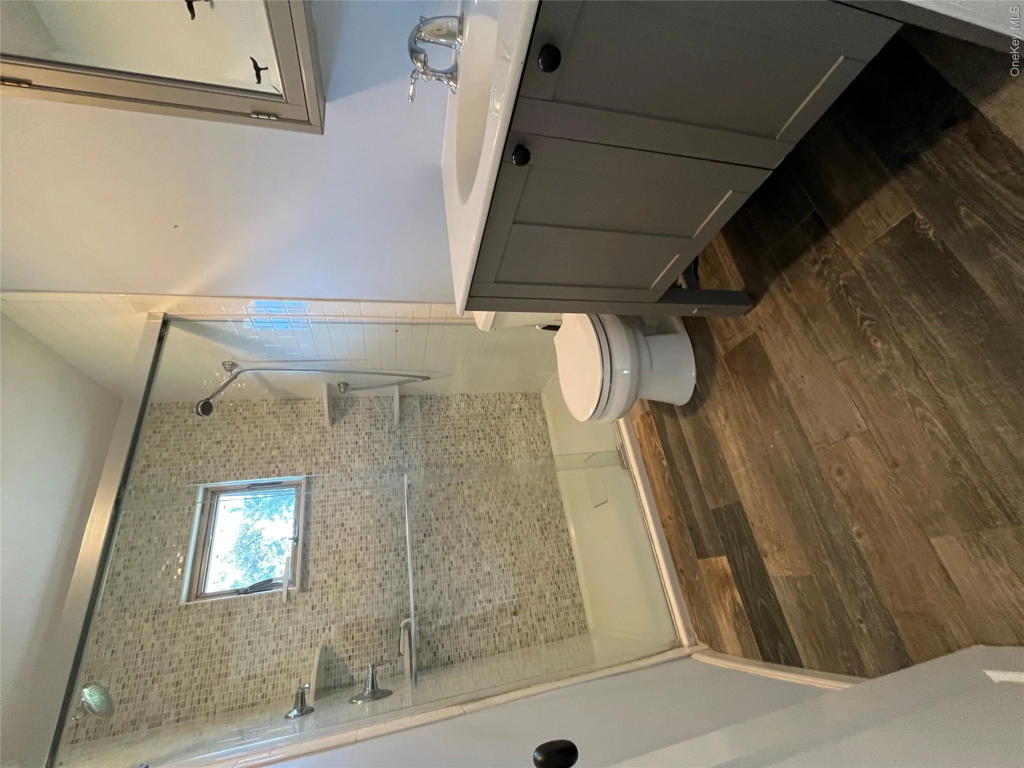 Remodeled beautiful Bathroom featuring wood-finish flooring, a frameless glass shower enclosure with mosaic tile surround, a built-in shower niche, and a vanity with a white countertop Remodeled beautiful Bathroom featuring wood-finish flooring, a frameless glass shower enclosure with mosaic tile surround, a built-in shower niche, and a vanity with a white countertop