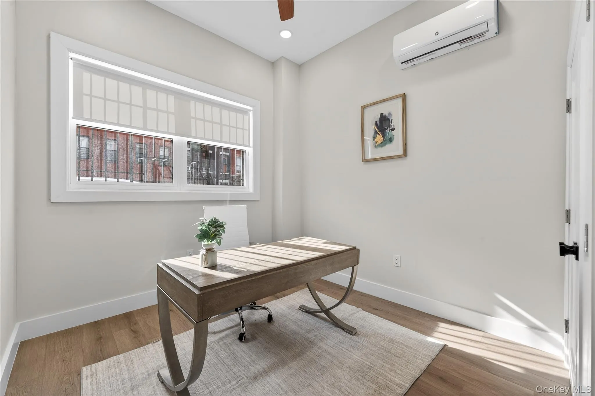 Bright room featuring a large window with roller shades, wood-finish flooring, and a wall-mounted air conditioning unit Bright room featuring a large window with roller shades, wood-finish flooring, and a wall-mounted air conditioning unit