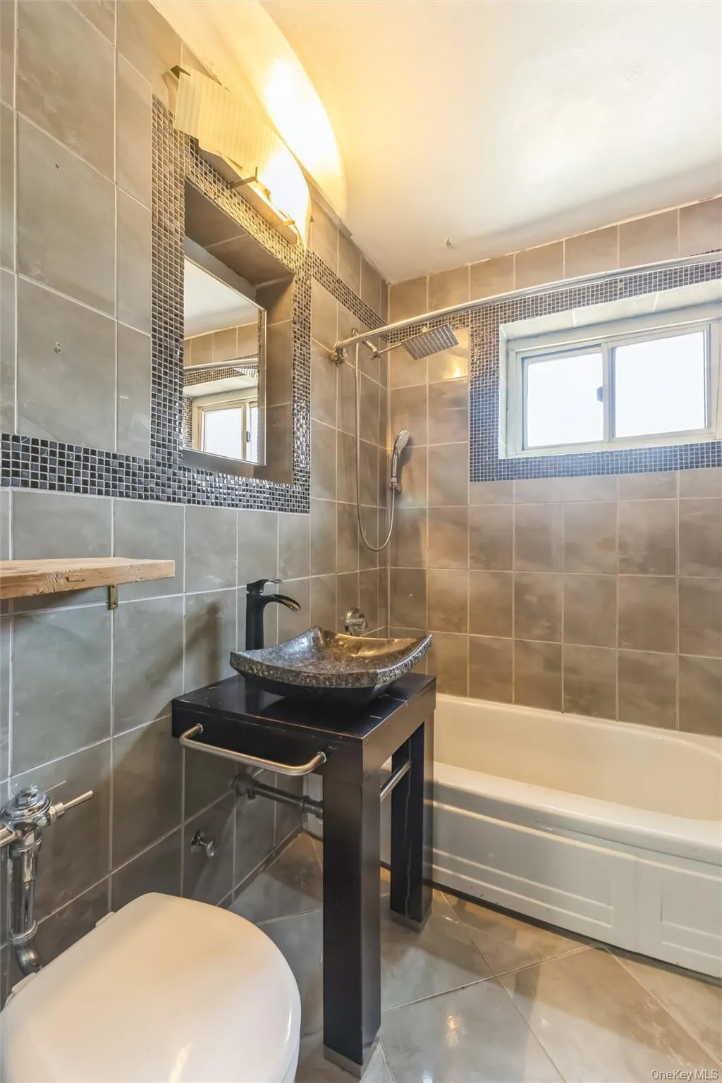 Bathroom featuring a stone vessel sink on a dark wood vanity, a full bathtub, a wall-mounted rain shower head, and tiled walls with decorative mosaic accents Bathroom featuring a stone vessel sink on a dark wood vanity, a full bathtub, a wall-mounted rain shower head, and tiled walls with decorative mosaic accents