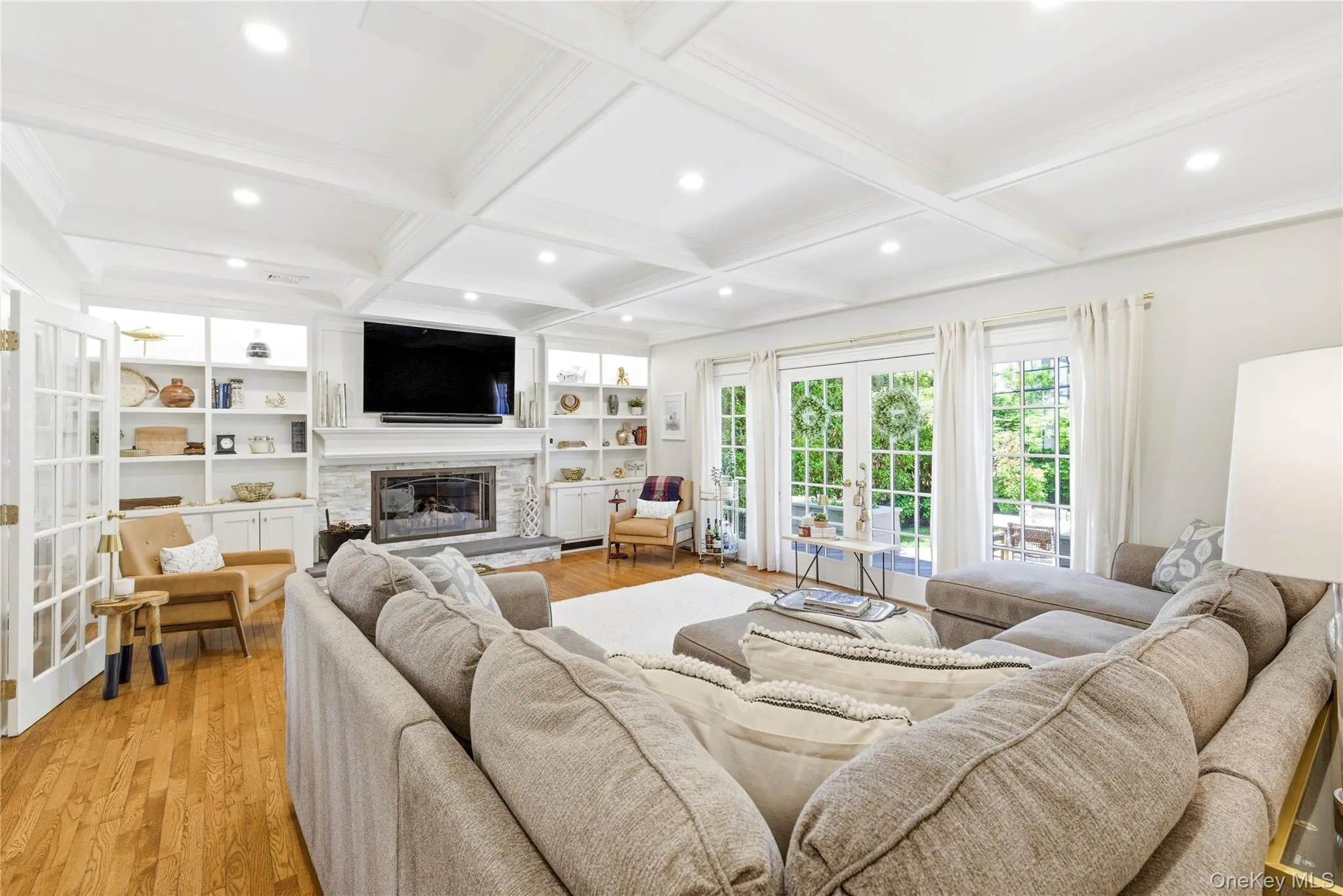 Living area featuring a coffered ceiling with recessed lighting, wood-finish flooring, a stone-faced fireplace with a mantel, and built-in shelving with cabinetry Living area featuring a coffered ceiling with recessed lighting, wood-finish flooring, a stone-faced fireplace with a mantel, and built-in shelving with cabinetry