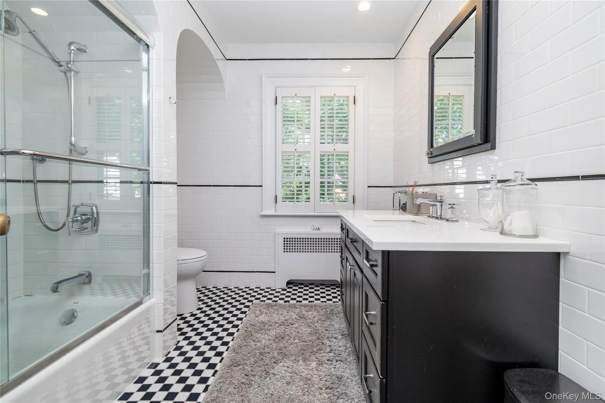 Bathroom featuring black and white checkerboard floor tiling, white subway wall tile, a dark wood vanity with a white countertop, a built-in tub with a sliding glass door, and a window with plantation shutters Bathroom featuring black and white checkerboard floor tiling, white subway wall tile, a dark wood vanity with a white countertop, a built-in tub with a sliding glass door, and a window with plantation shutters