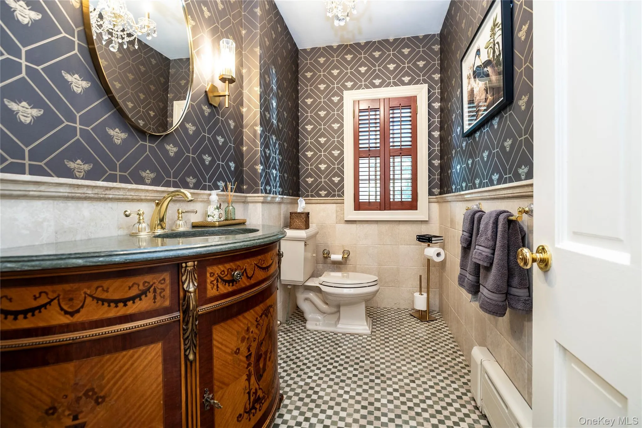 Bathroom featuring a patterned wall covering, wood-finish vanity with a gold-tone faucet, stone countertop, checkered floor tile, and a window with wood shutters Bathroom featuring a patterned wall covering, wood-finish vanity with a gold-tone faucet, stone countertop, checkered floor tile, and a window with wood shutters