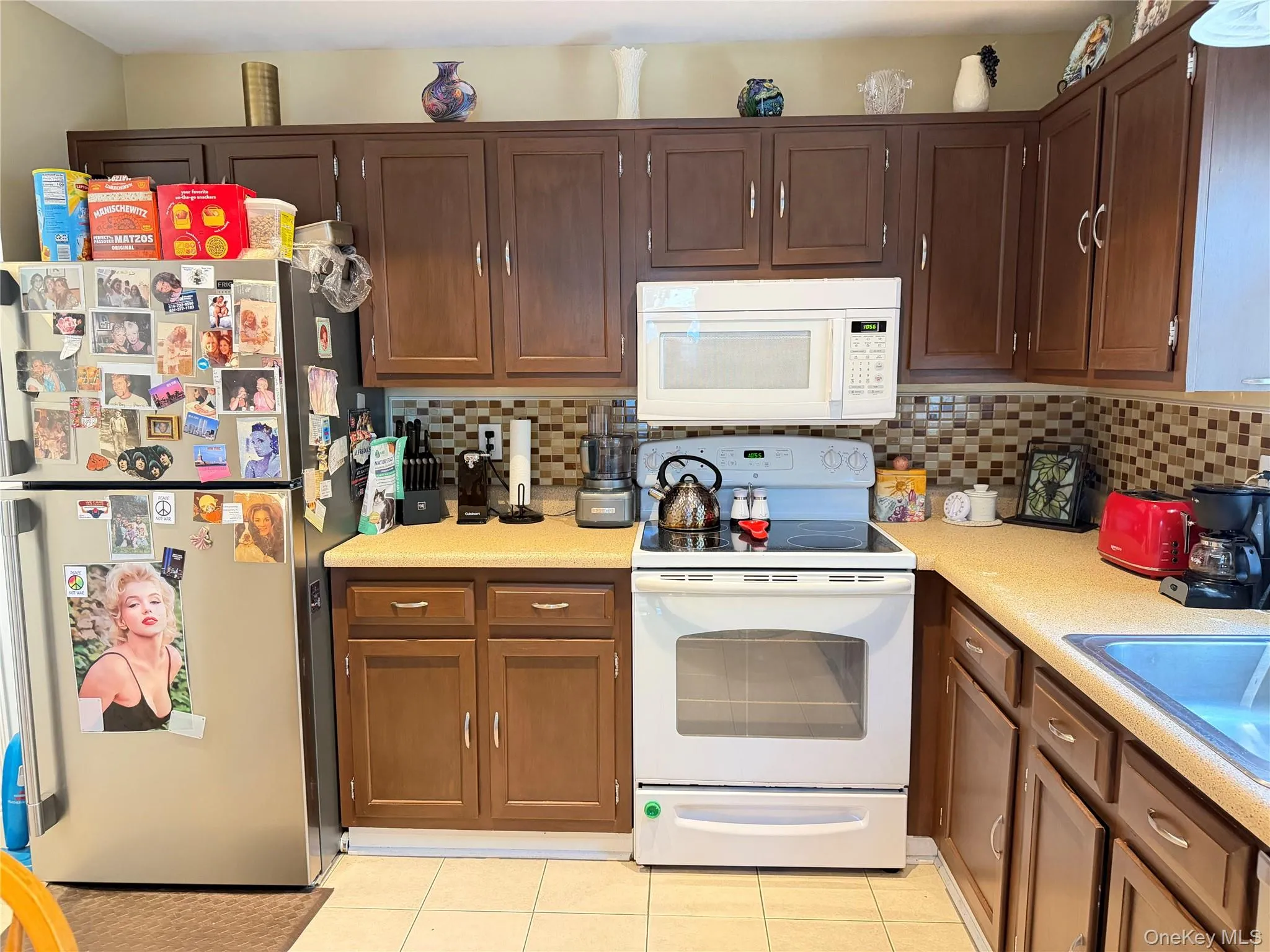 Kitchen featuring dark wood-finish cabinetry, a mosaic tile backsplash, and a stainless steel dishwasher with sliding doors leading to patio Kitchen featuring dark wood-finish cabinetry, a mosaic tile backsplash, and a stainless steel dishwasher with sliding doors leading to patio