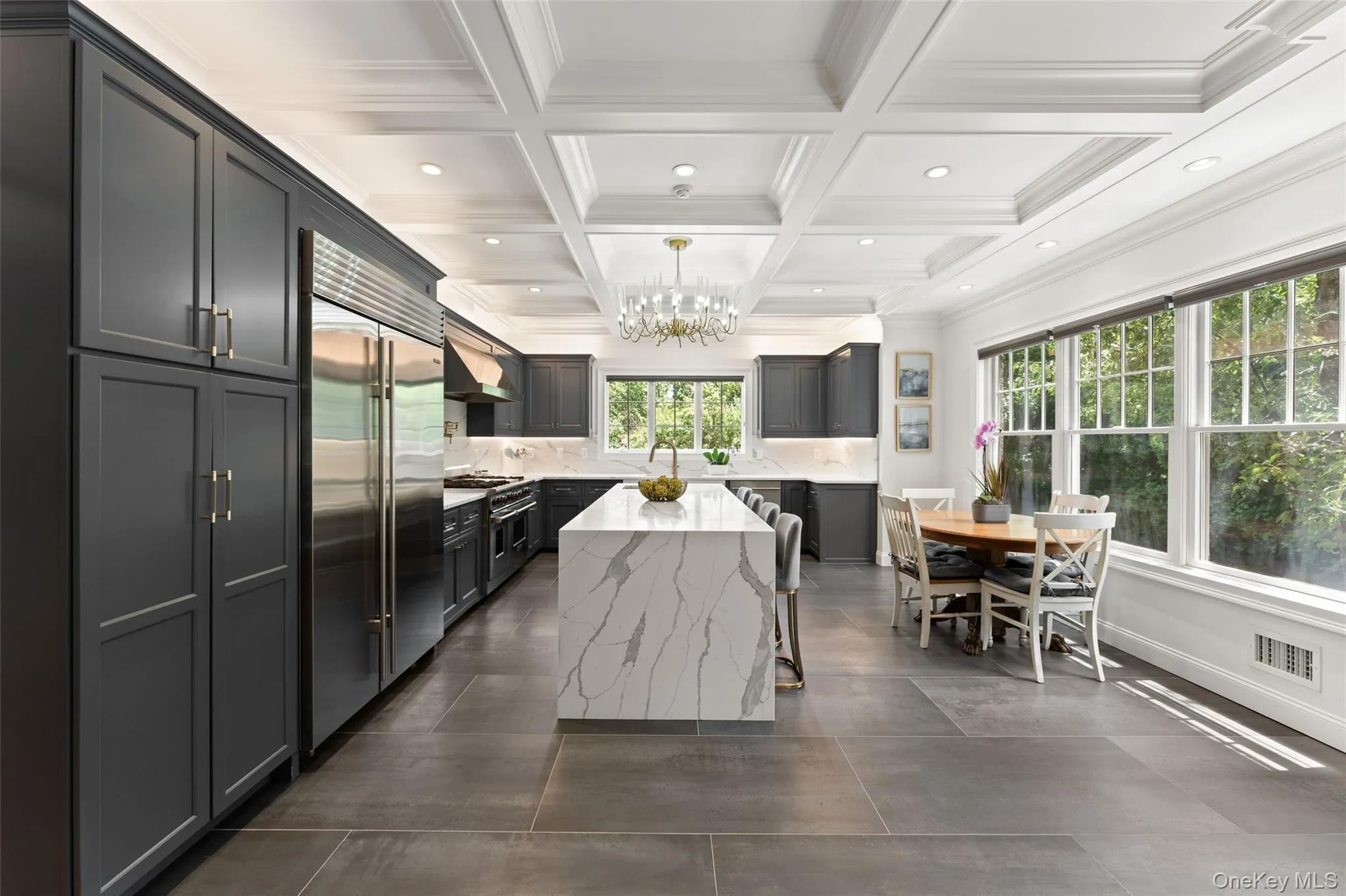 Gourmet kitchen featuring a large waterfall island with seating, stainless steel appliances, dark shaker cabinetry, a coffered ceiling with recessed lighting, and large windows providing abundant natural light Gourmet kitchen featuring a large waterfall island with seating, stainless steel appliances, dark shaker cabinetry, a coffered ceiling with recessed lighting, and large windows providing abundant natural light
