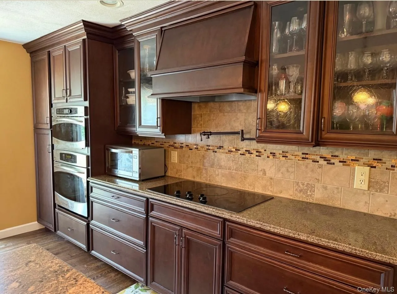 Kitchen featuring rich wood cabinetry with crown molding, double stainless steel wall ovens, a black cooktop, a neutral tiled backsplash with an accent border, and speckled countertops Digitally removed personal items Kitchen featuring rich wood cabinetry with crown molding, double stainless steel wall ovens, a black cooktop, a neutral tiled backsplash with an accent border, and speckled countertops Digitally removed personal items