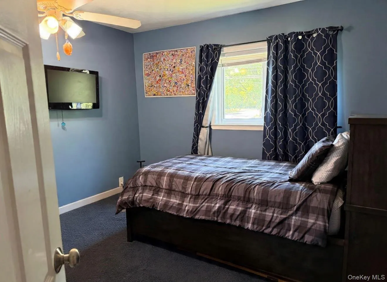 Bedroom featuring blue wall paint, dark carpeting, and a window with patterned curtains Digitally removed personal items Bedroom featuring blue wall paint, dark carpeting, and a window with patterned curtains Digitally removed personal items