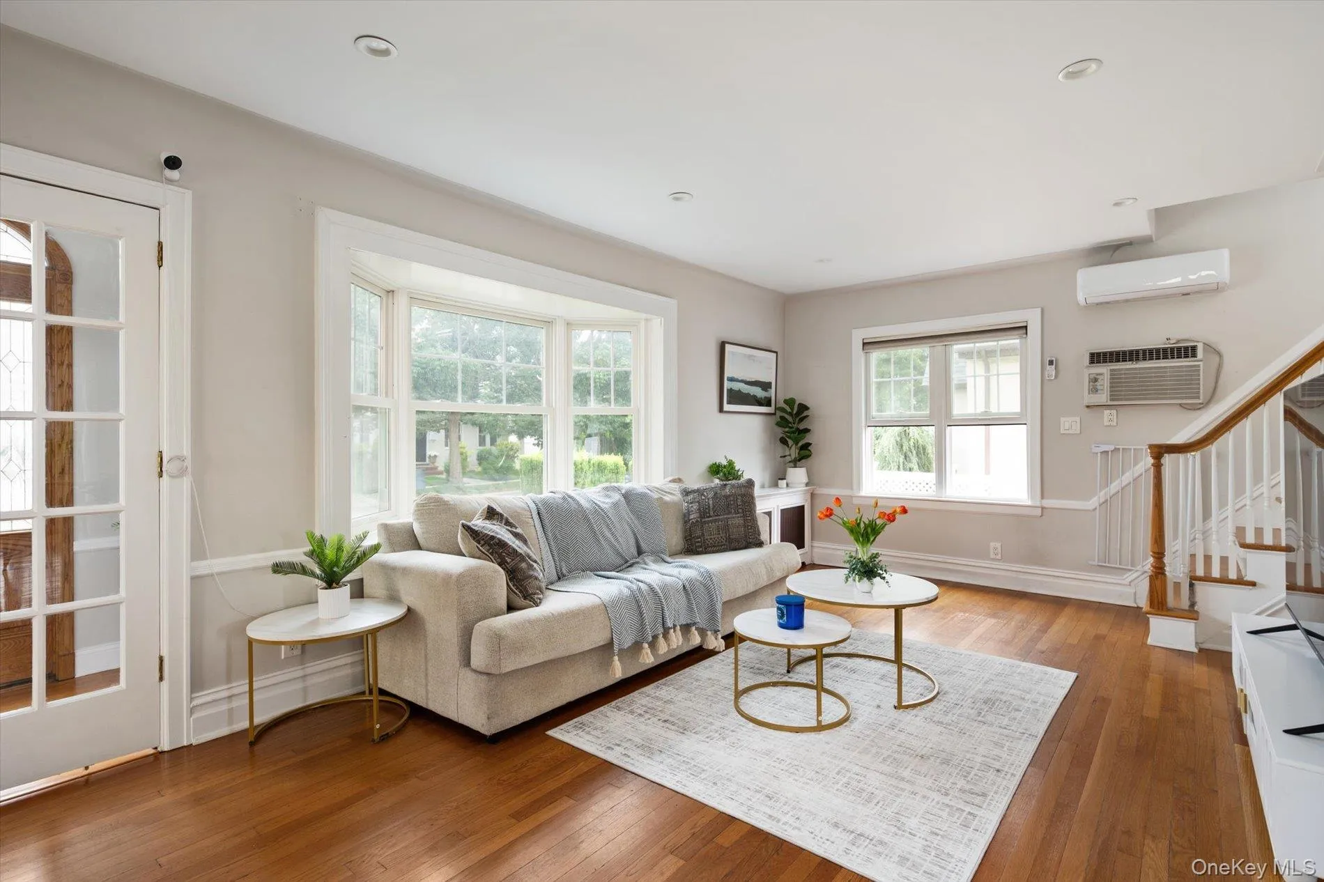 Living area featuring wood-finish flooring, a bay window with multiple panes, a white painted staircase with wood banister, recessed lighting, and a glass-paneled door Living area featuring wood-finish flooring, a bay window with multiple panes, a white painted staircase with wood banister, recessed lighting, and a glass-paneled door