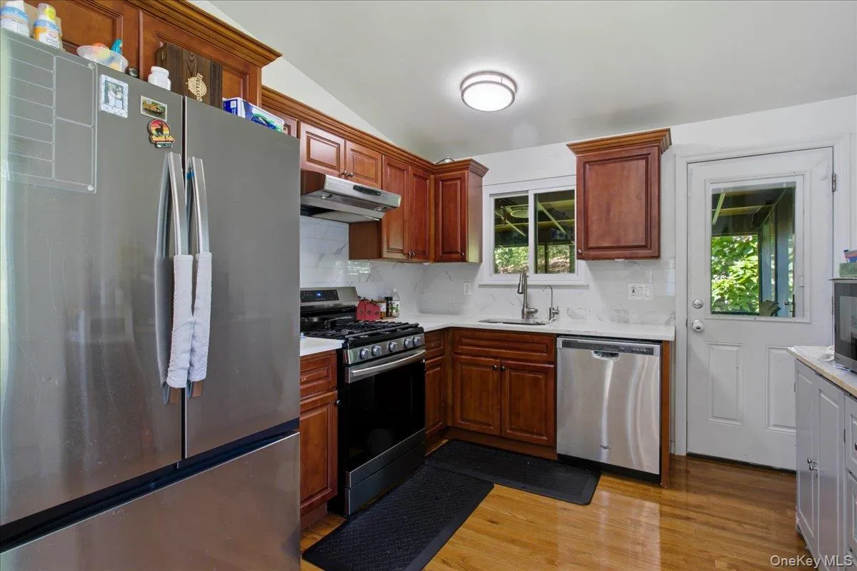 Kitchen featuring wood-finish cabinetry, stainless steel appliances, white countertops, a tile backsplash, and wood-finish flooring Kitchen featuring wood-finish cabinetry, stainless steel appliances, white countertops, a tile backsplash, and wood-finish flooring