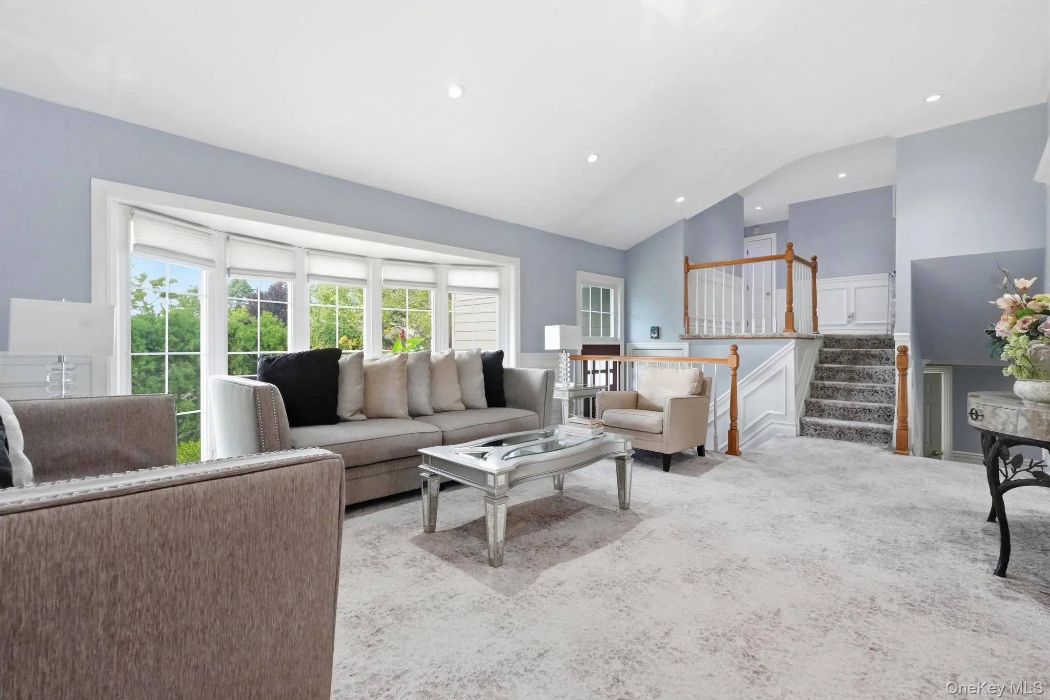 Spacious living area featuring a large bay window with outdoor views, recessed lighting, light-toned carpeting, and a staircase with wood railings Spacious living area featuring a large bay window with outdoor views, recessed lighting, light-toned carpeting, and a staircase with wood railings
