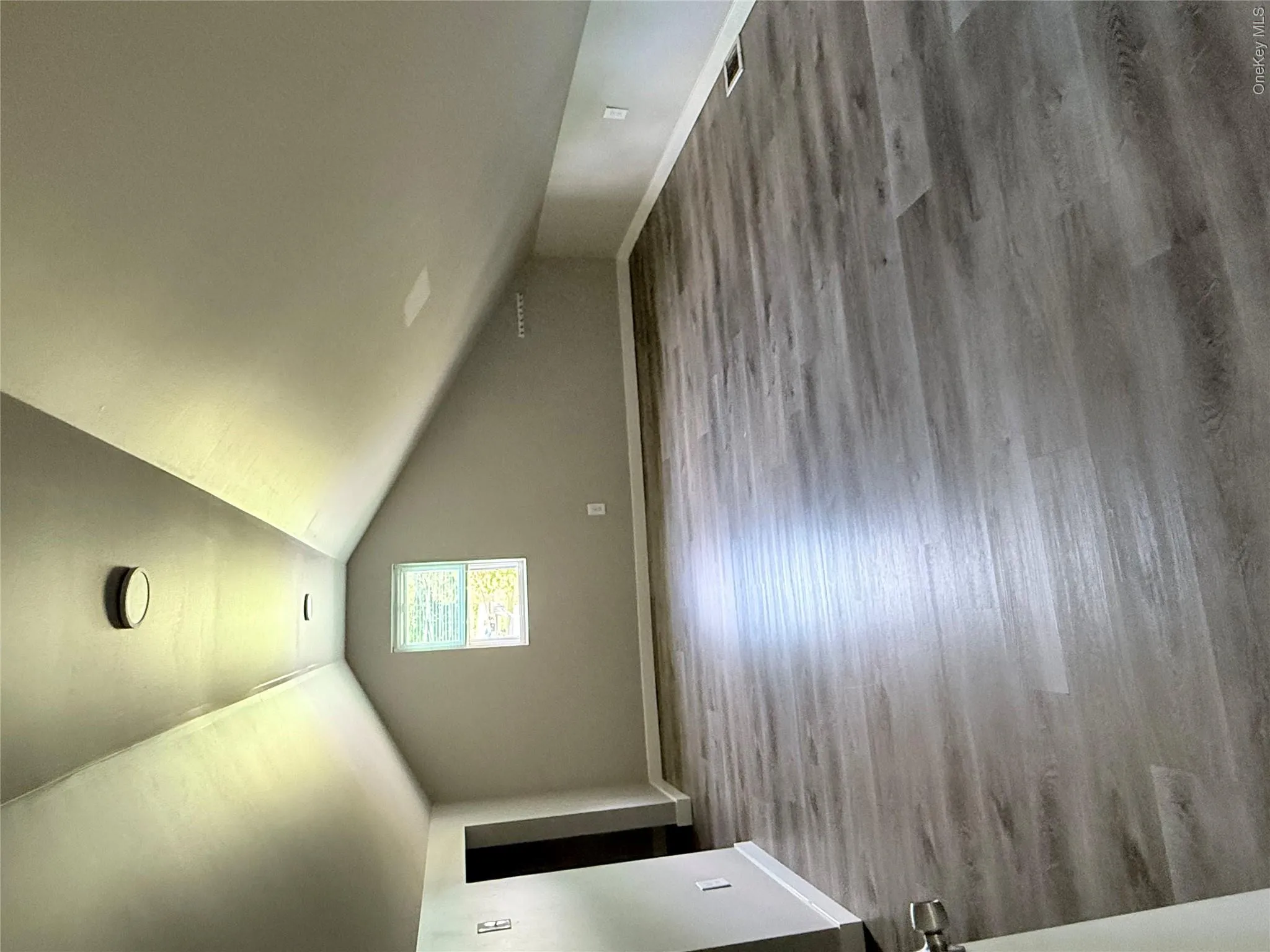 Spacious room with sloped ceilings, modern wood-finish flooring, a single window, and flush-mount lighting fixtures Spacious room with sloped ceilings, modern wood-finish flooring, a single window, and flush-mount lighting fixtures