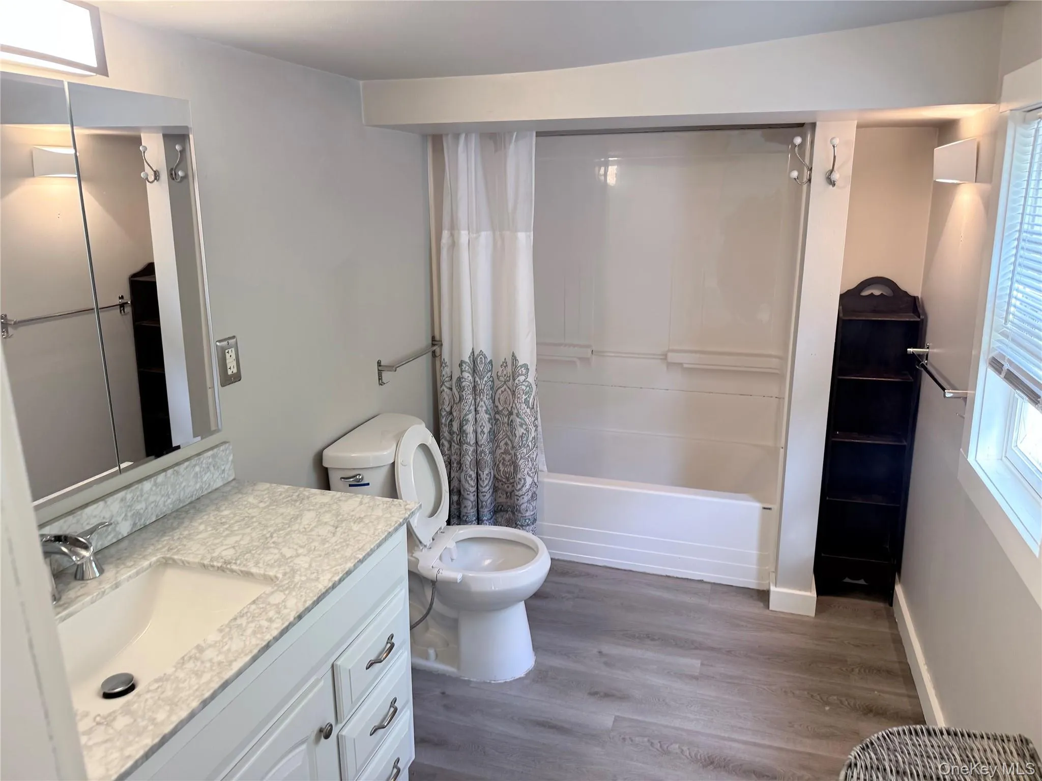 Bathroom featuring a vanity with an integrated sink and light-colored countertop, wood-finish flooring, a white tub-shower combination, and a window with horizontal blinds Bathroom featuring a vanity with an integrated sink and light-colored countertop, wood-finish flooring, a white tub-shower combination, and a window with horizontal blinds