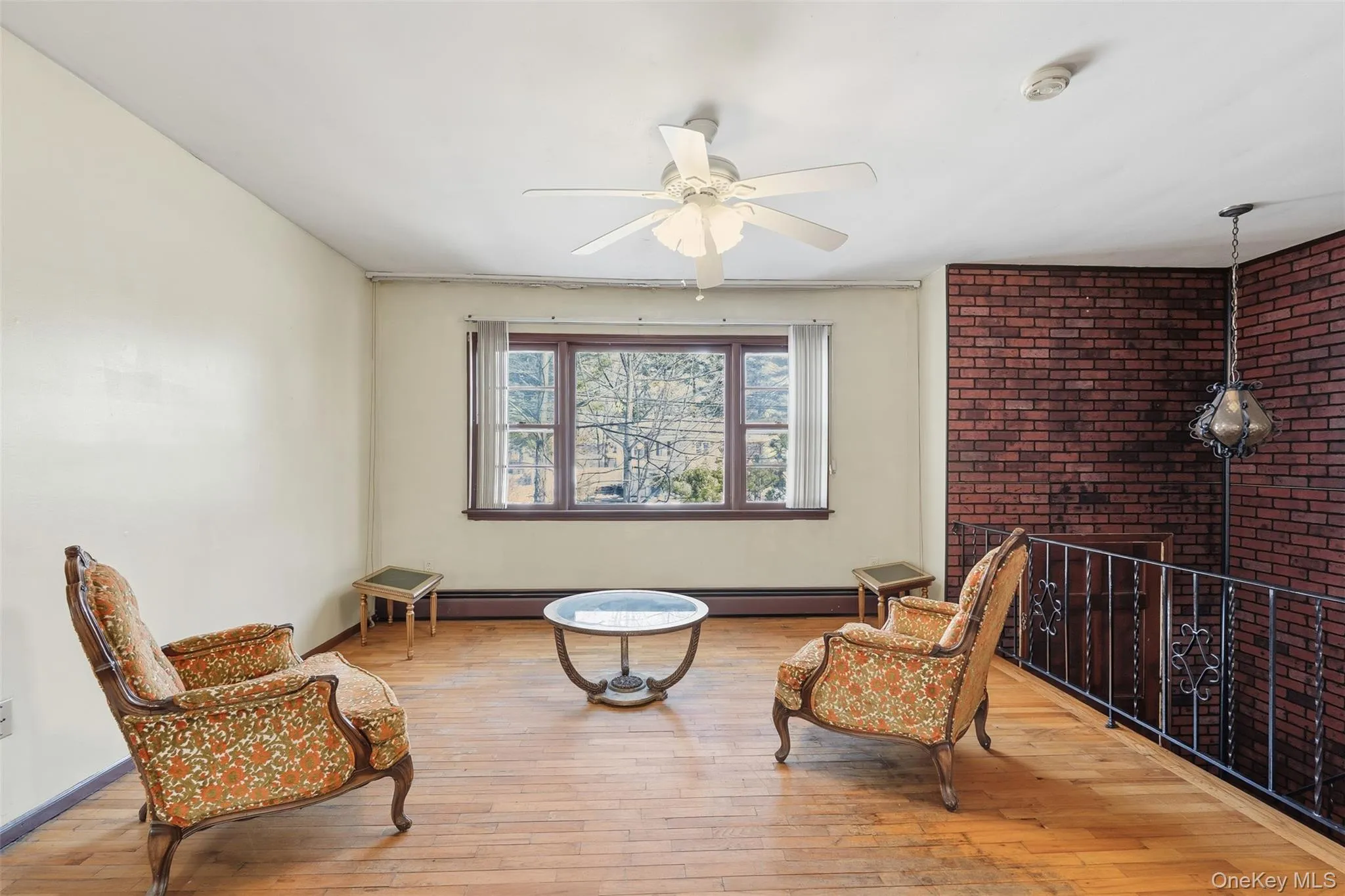 Expansive room featuring wood-finish flooring, a red brick accent wall, and a multi-pane window Expansive room featuring wood-finish flooring, a red brick accent wall, and a multi-pane window