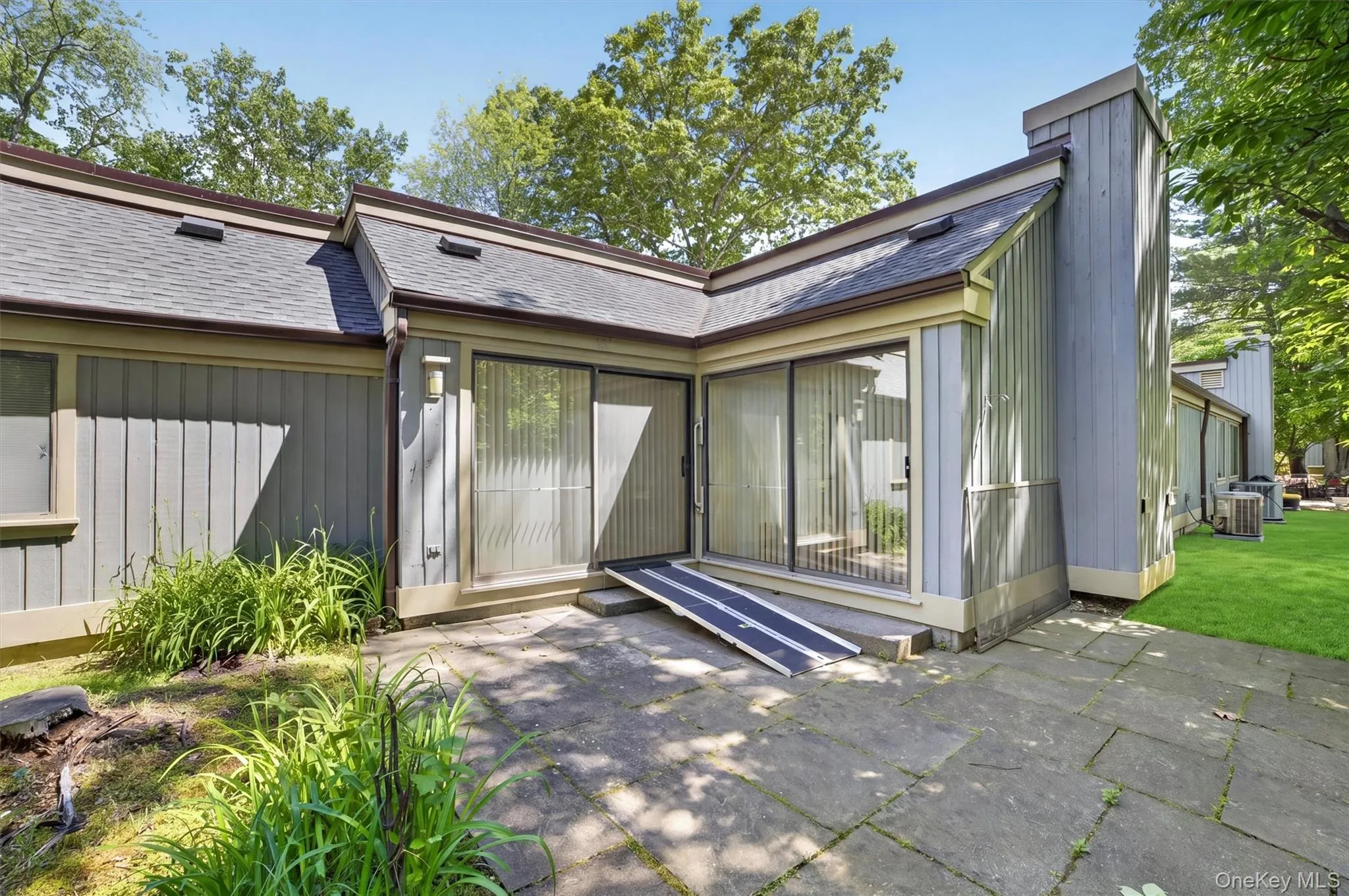 Paved stone patio featuring two sliding glass doors, a wood-sided exterior, a shingled roof, and a permanent access ramp Paved stone patio featuring two sliding glass doors, a wood-sided exterior, a shingled roof, and a permanent access ramp