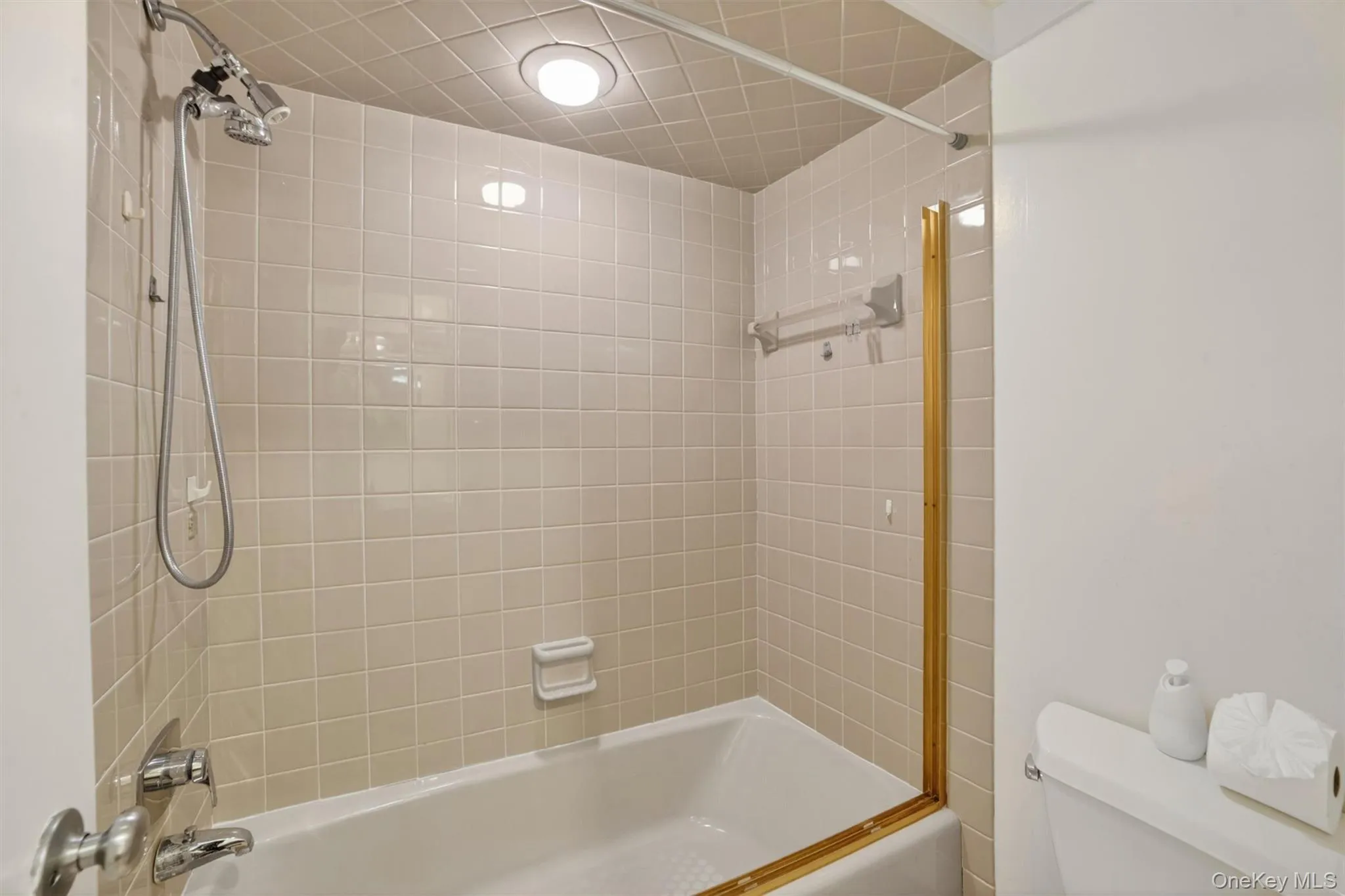 Tub and shower combination featuring neutral square tile surround, chrome fixtures with handheld showerhead, and a recessed ceiling light Tub and shower combination featuring neutral square tile surround, chrome fixtures with handheld showerhead, and a recessed ceiling light