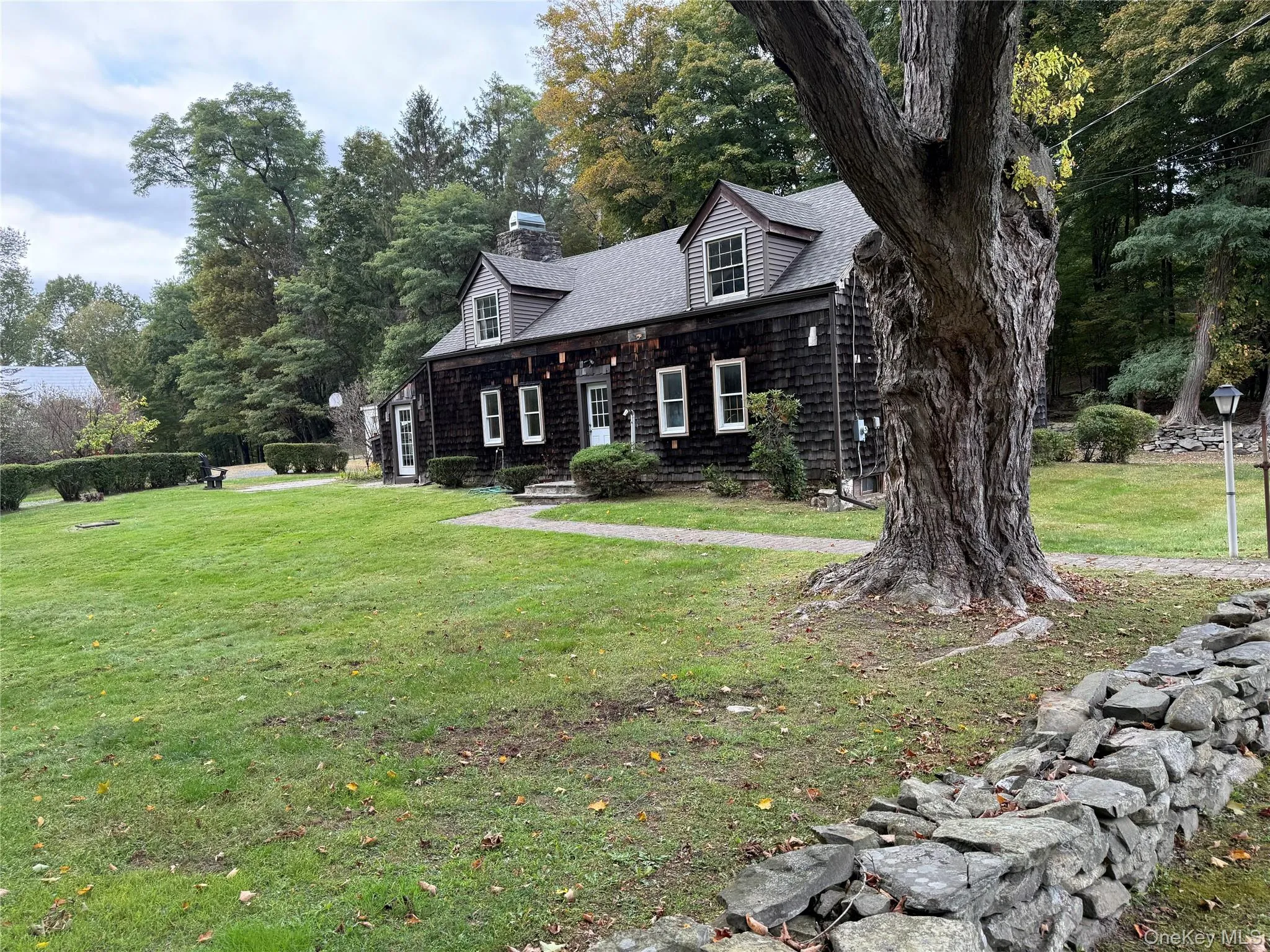 New england style home featuring a front yard, a chimney, view of wooded area, and roof with shingles New england style home featuring a front yard, a chimney, view of wooded area, and roof with shingles