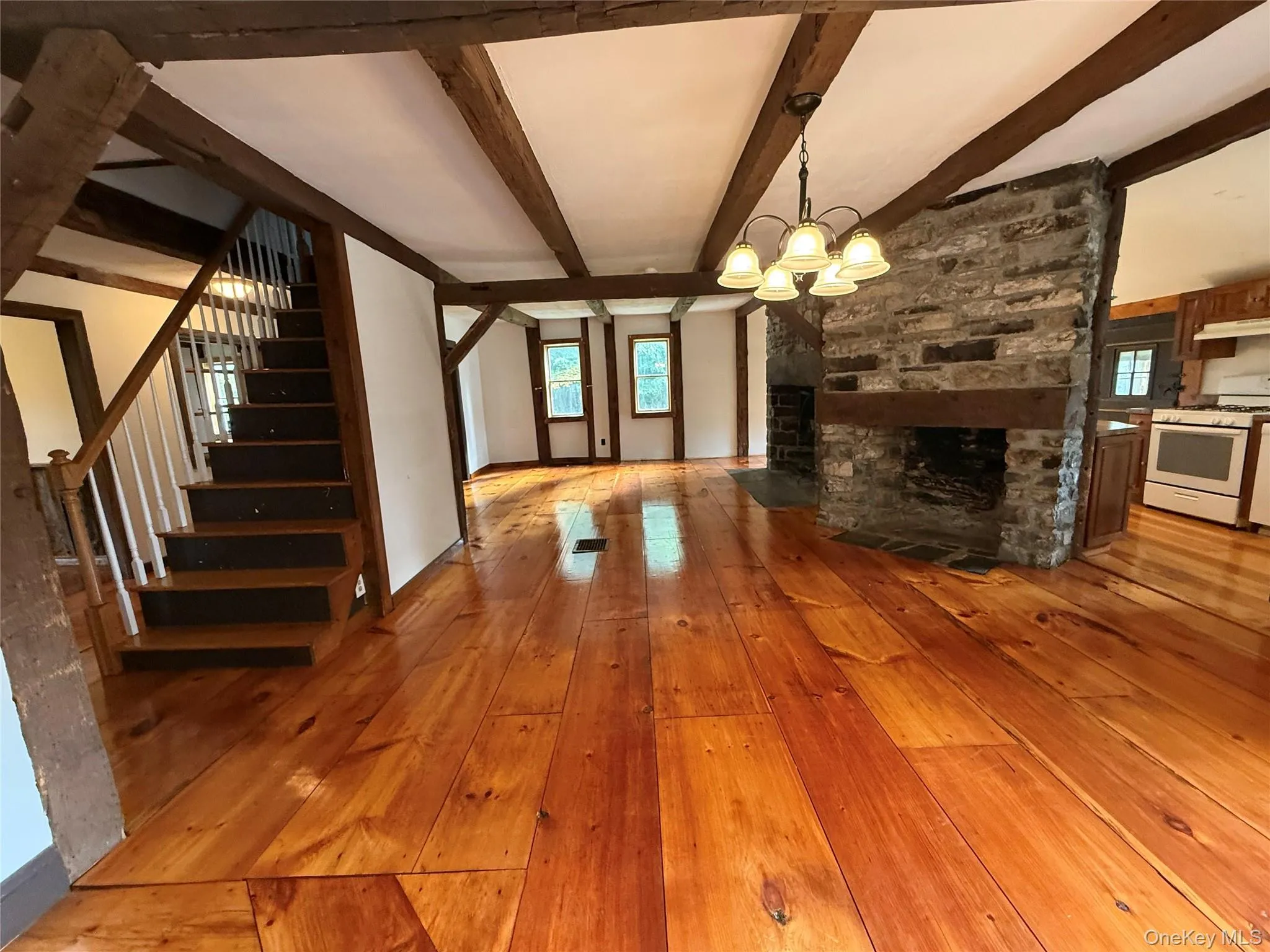 Unfurnished living room with light wood finished floors, a stone fireplace, beamed ceiling, stairs, and a chandelier Unfurnished living room with light wood finished floors, a stone fireplace, beamed ceiling, stairs, and a chandelier