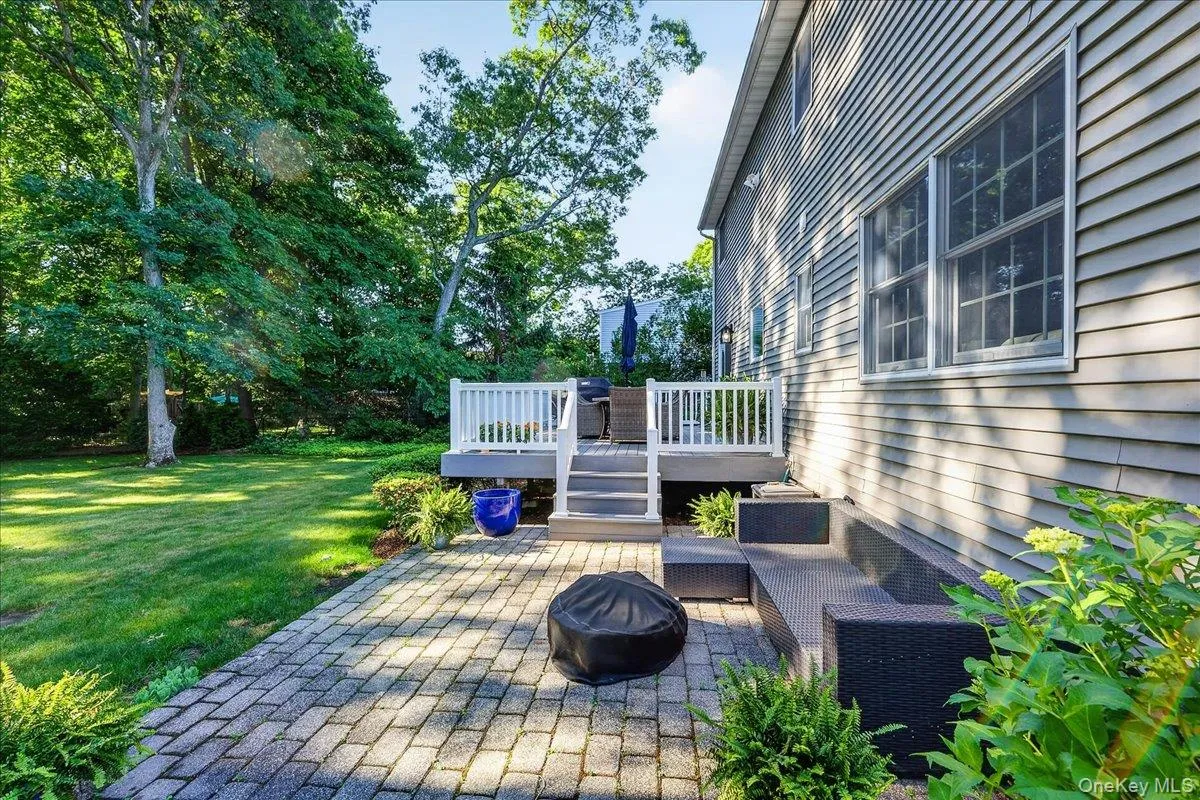 Expansive backyard featuring a paver patio with built-in seating, a raised deck with white railings, and a manicured lawn framed by mature trees Expansive backyard featuring a paver patio with built-in seating, a raised deck with white railings, and a manicured lawn framed by mature trees