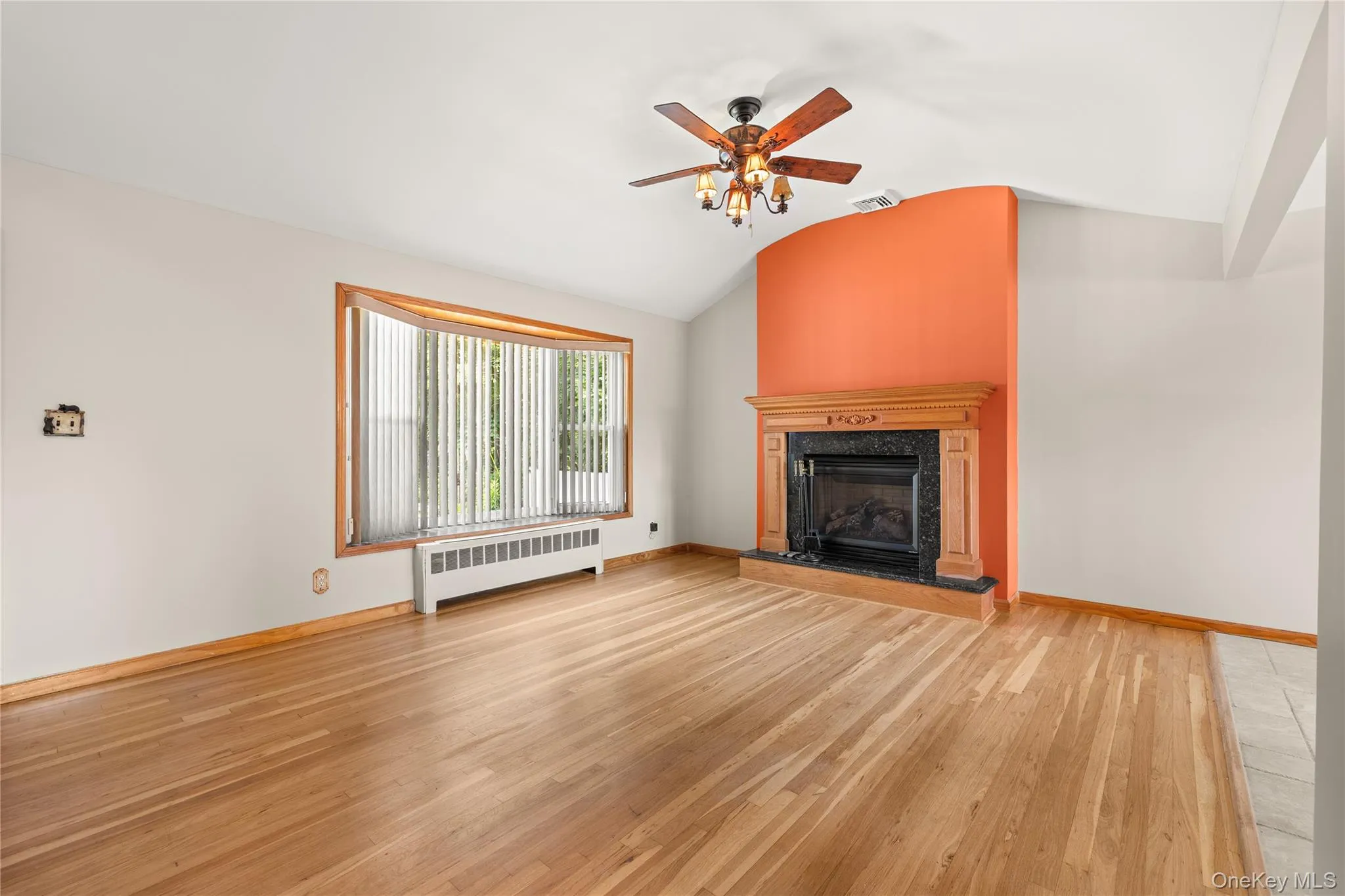 Vaulted ceiling living space featuring wood-finish flooring, a wood-mantel fireplace with a dark stone surround, a bay window, and a ceiling fan with integrated lighting Vaulted ceiling living space featuring wood-finish flooring, a wood-mantel fireplace with a dark stone surround, a bay window, and a ceiling fan with integrated lighting