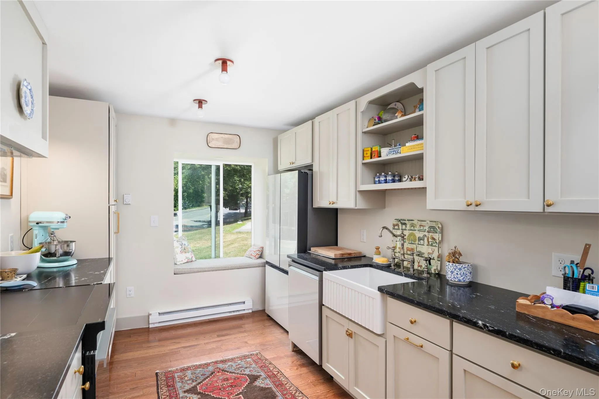 Kitchen featuring light-toned cabinetry, dark countertops, wood-finish flooring, a farmhouse-style sink, and a window seat overlooking exterior greenery Kitchen featuring light-toned cabinetry, dark countertops, wood-finish flooring, a farmhouse-style sink, and a window seat overlooking exterior greenery