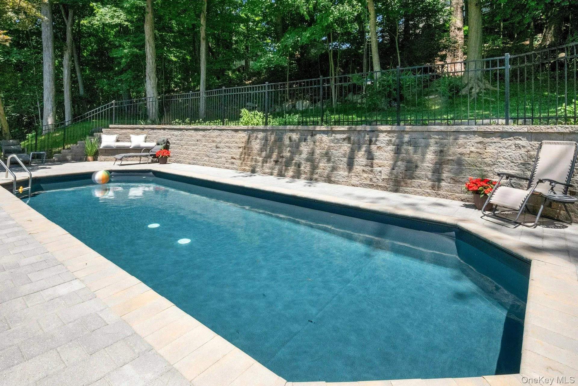 Reverse view of pool with lounge chairs and retaining wall behind with fencing above. Reverse view of pool with lounge chairs and retaining wall behind with fencing above.