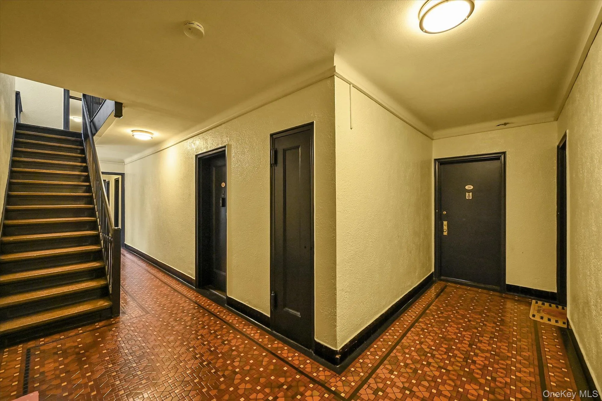 Corridor featuring wood-finish flooring with a patterned inlay, a dark wood-finish staircase with matching handrails, and multiple dark-paneled entry doors. Corridor featuring wood-finish flooring with a patterned inlay, a dark wood-finish staircase with matching handrails, and multiple dark-paneled entry doors.