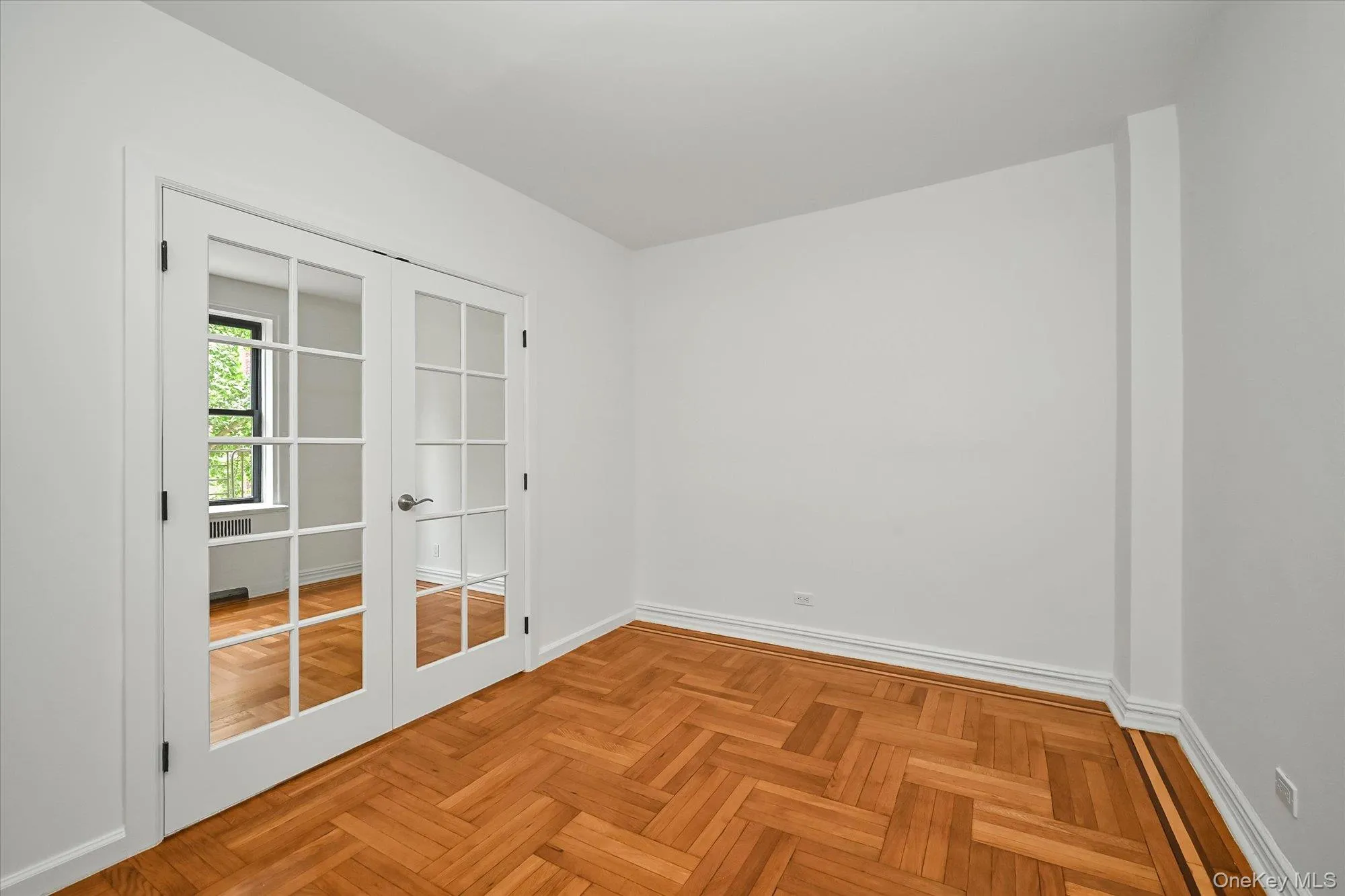 Room featuring white French doors with divided lites, herringbone wood-finish flooring, white baseboards, and painted white walls. Room featuring white French doors with divided lites, herringbone wood-finish flooring, white baseboards, and painted white walls.
