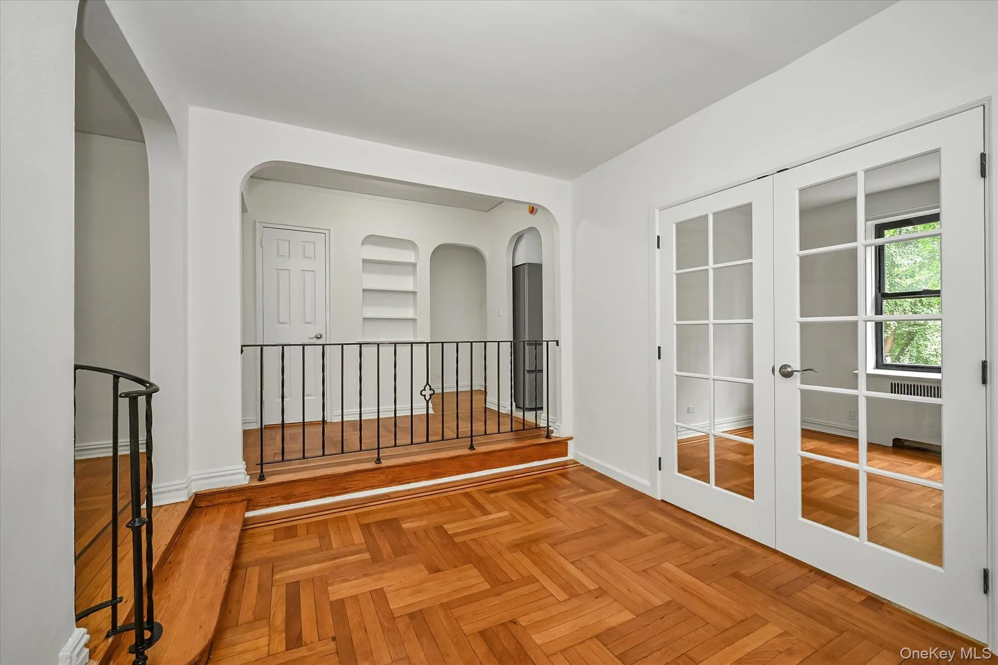 Herringbone wood-finish flooring, white French doors with glass panels, arched doorways, built-in shelving, and a decorative wrought iron railing. Herringbone wood-finish flooring, white French doors with glass panels, arched doorways, built-in shelving, and a decorative wrought iron railing.