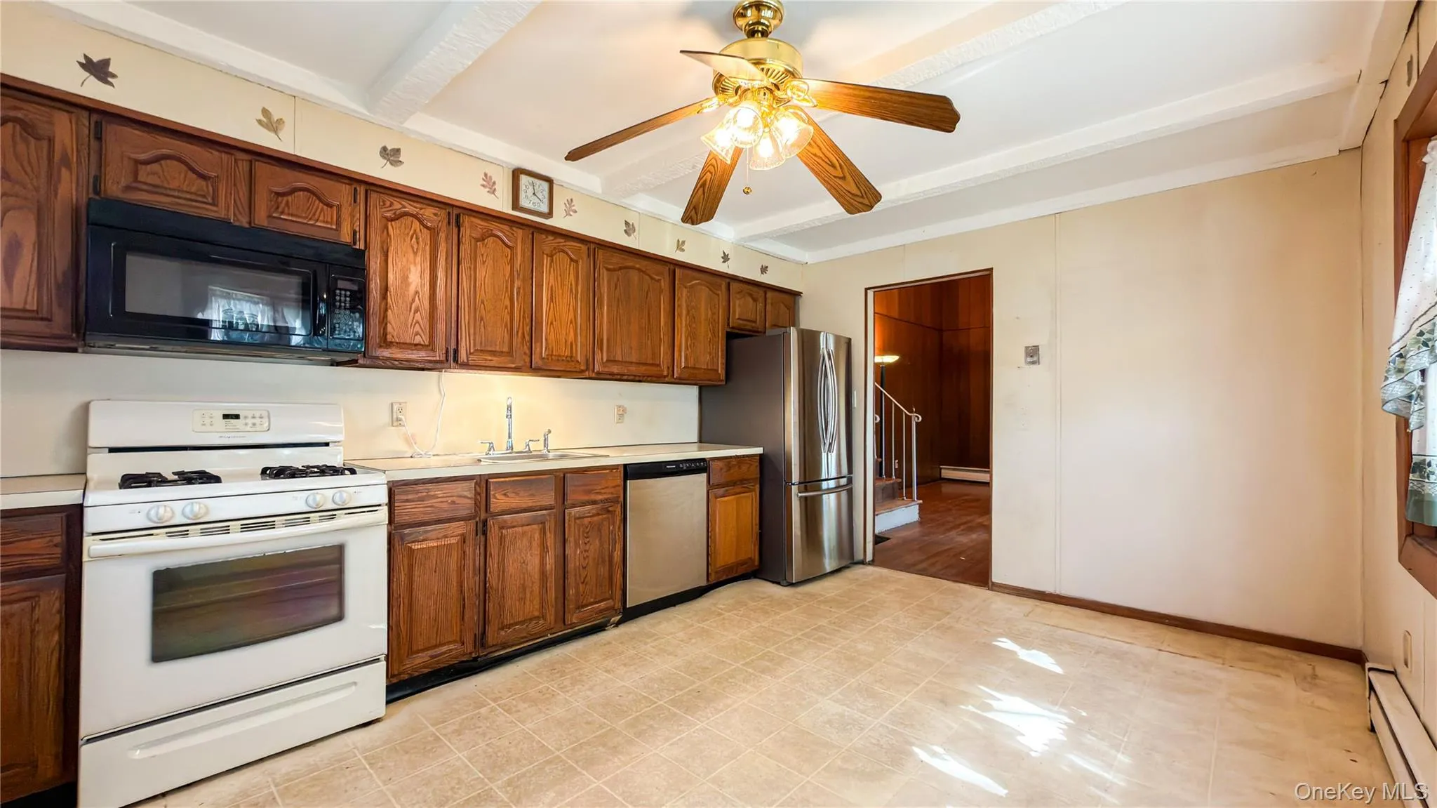 Eat-in-kitchen with wood cabinets, stainless steel refrigerator and dishwasher, white gas stove, black microwave with white painted beam ceiling, ceiling fan, linoleum floors leading into living room Eat-in-kitchen with wood cabinets, stainless steel refrigerator and dishwasher, white gas stove, black microwave with white painted beam ceiling, ceiling fan, linoleum floors leading into living room