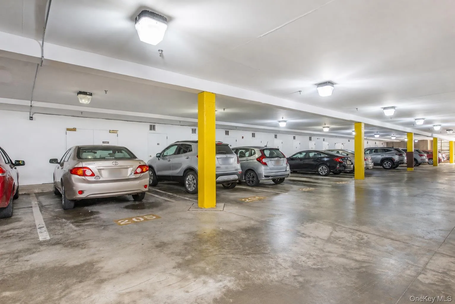 Underground parking garage featuring concrete flooring, white walls, yellow support columns, and overhead lighting fixtures Underground parking garage featuring concrete flooring, white walls, yellow support columns, and overhead lighting fixtures