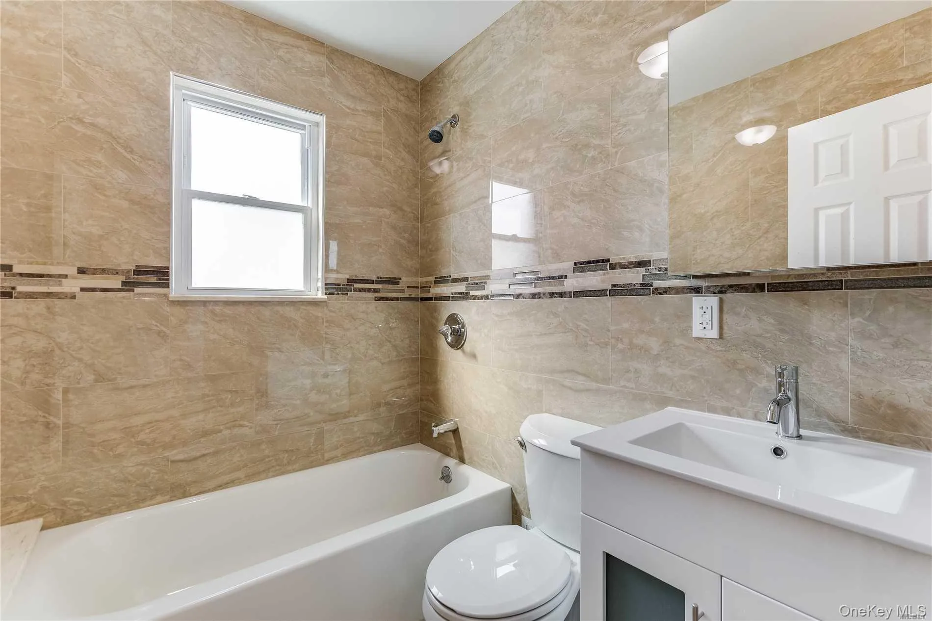 Bathroom featuring full-height stone-look tile, decorative accent tile band, white single-hung window, white bathtub, and vanity with an integrated sink Bathroom featuring full-height stone-look tile, decorative accent tile band, white single-hung window, white bathtub, and vanity with an integrated sink