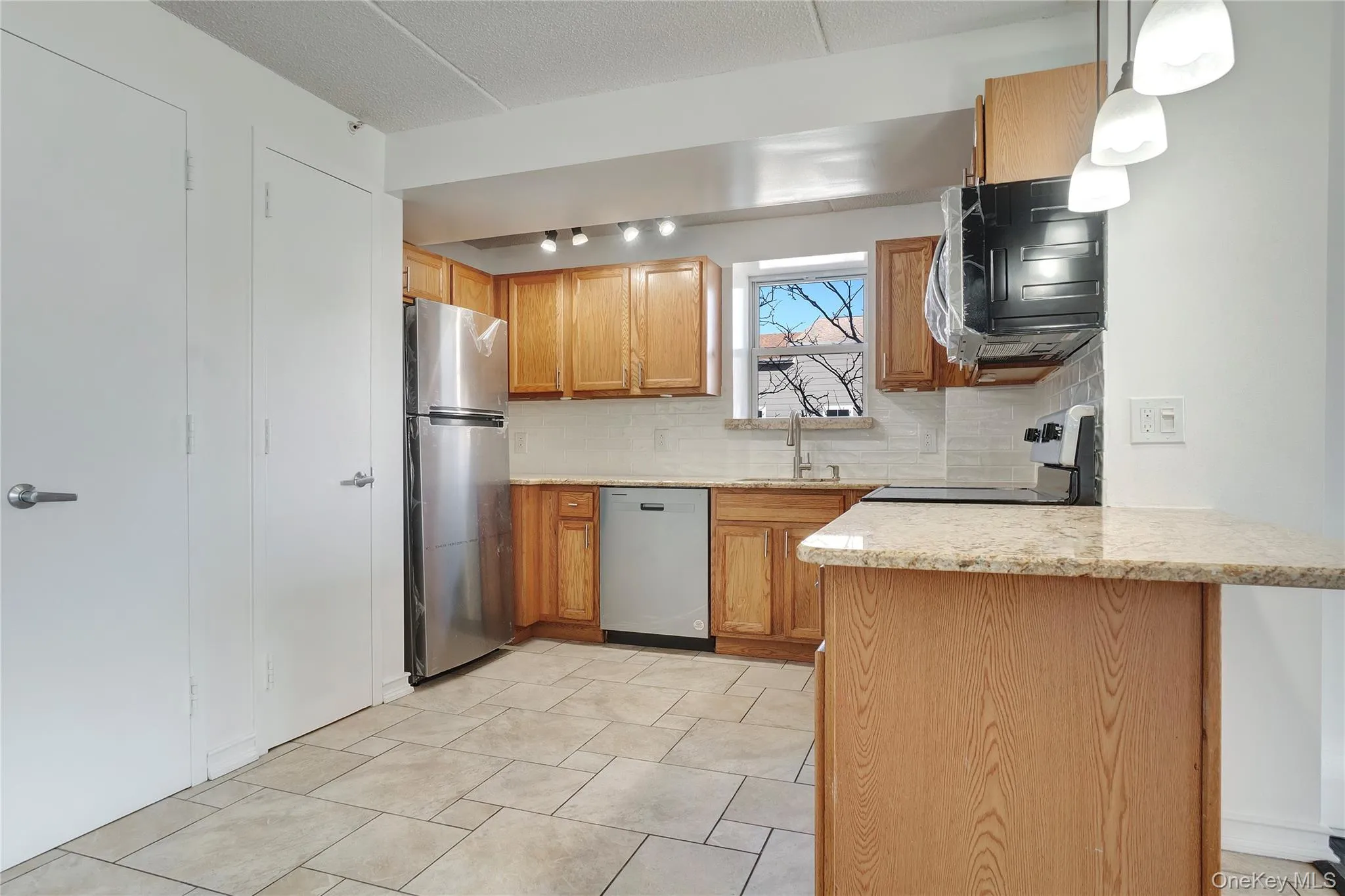 Kitchen featuring decorative backsplash, appliances with stainless steel finishes, a peninsula, brown cabinetry, and light stone countertops Kitchen featuring decorative backsplash, appliances with stainless steel finishes, a peninsula, brown cabinetry, and light stone countertops