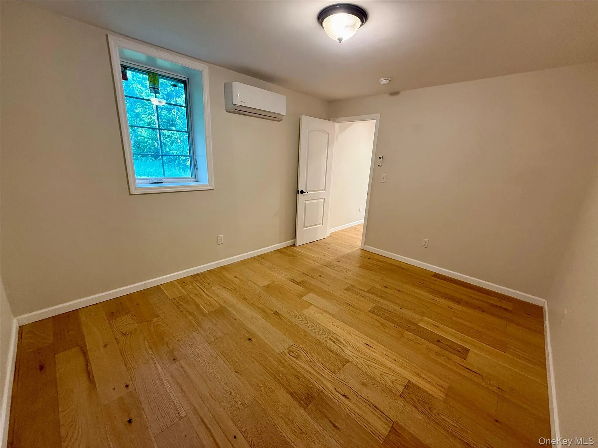 Hardwood flooring throughout, featuring a window with white trim, a wall-mounted mini-split AC unit, and a flush-mount ceiling light Hardwood flooring throughout, featuring a window with white trim, a wall-mounted mini-split AC unit, and a flush-mount ceiling light