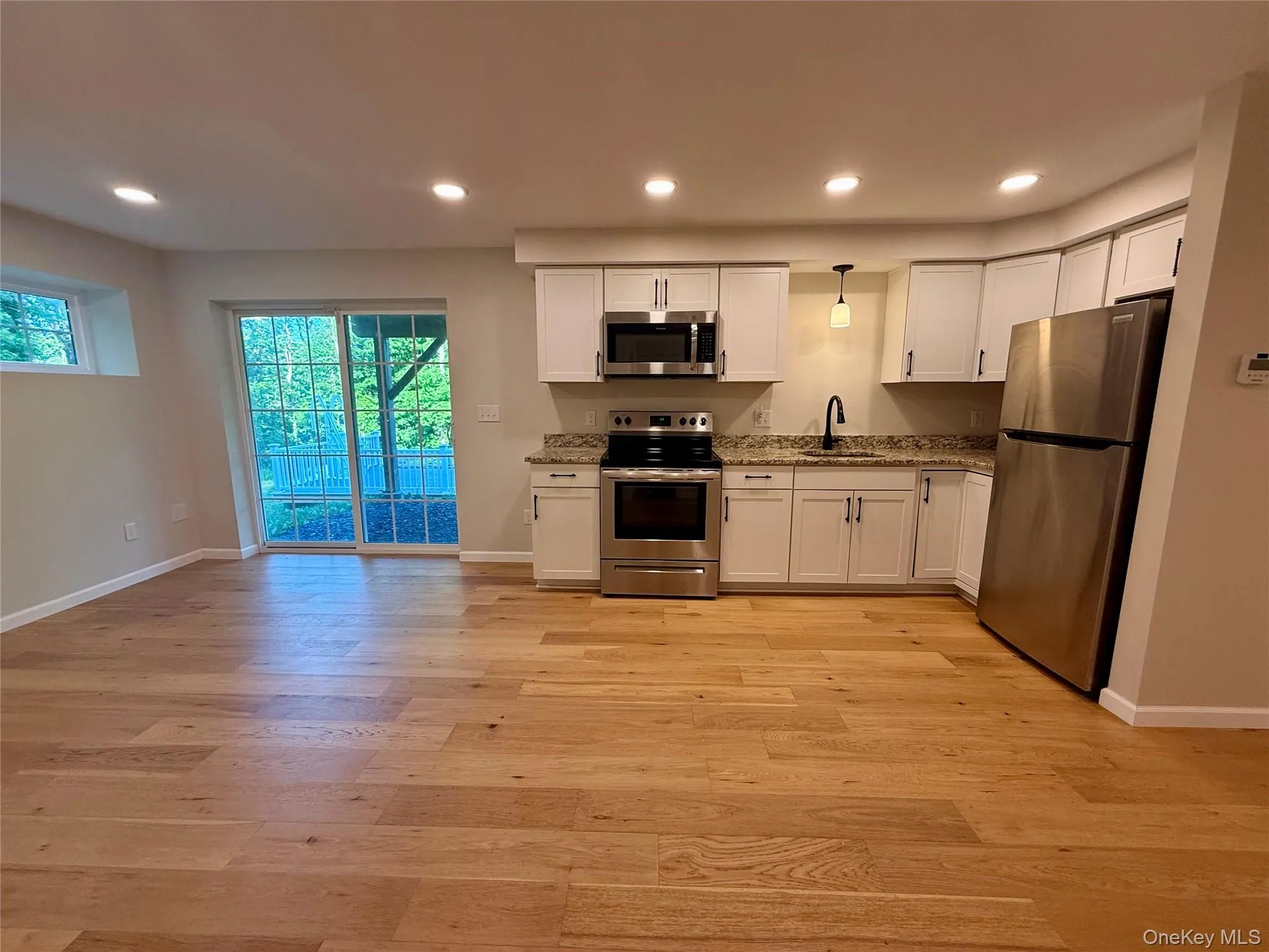 Open-concept space featuring wood-finish flooring, a kitchen with white cabinetry, stainless steel appliances, and granite countertops, recessed lighting, and a glass sliding door Open-concept space featuring wood-finish flooring, a kitchen with white cabinetry, stainless steel appliances, and granite countertops, recessed lighting, and a glass sliding door