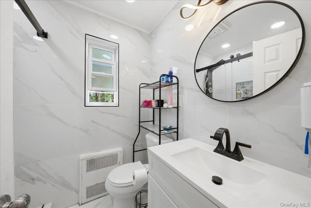 Bathroom featuring large format white tiled walls, a white vanity with integrated sink, black hardware throughout, an oval mirror, and a window Bathroom featuring large format white tiled walls, a white vanity with integrated sink, black hardware throughout, an oval mirror, and a window