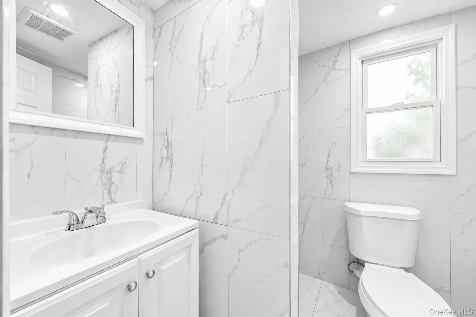 Bathroom featuring large format marble-look tile walls and flooring, a white vanity with an integrated sink, polished chrome faucet, and a recessed ceiling light Bathroom featuring large format marble-look tile walls and flooring, a white vanity with an integrated sink, polished chrome faucet, and a recessed ceiling light
