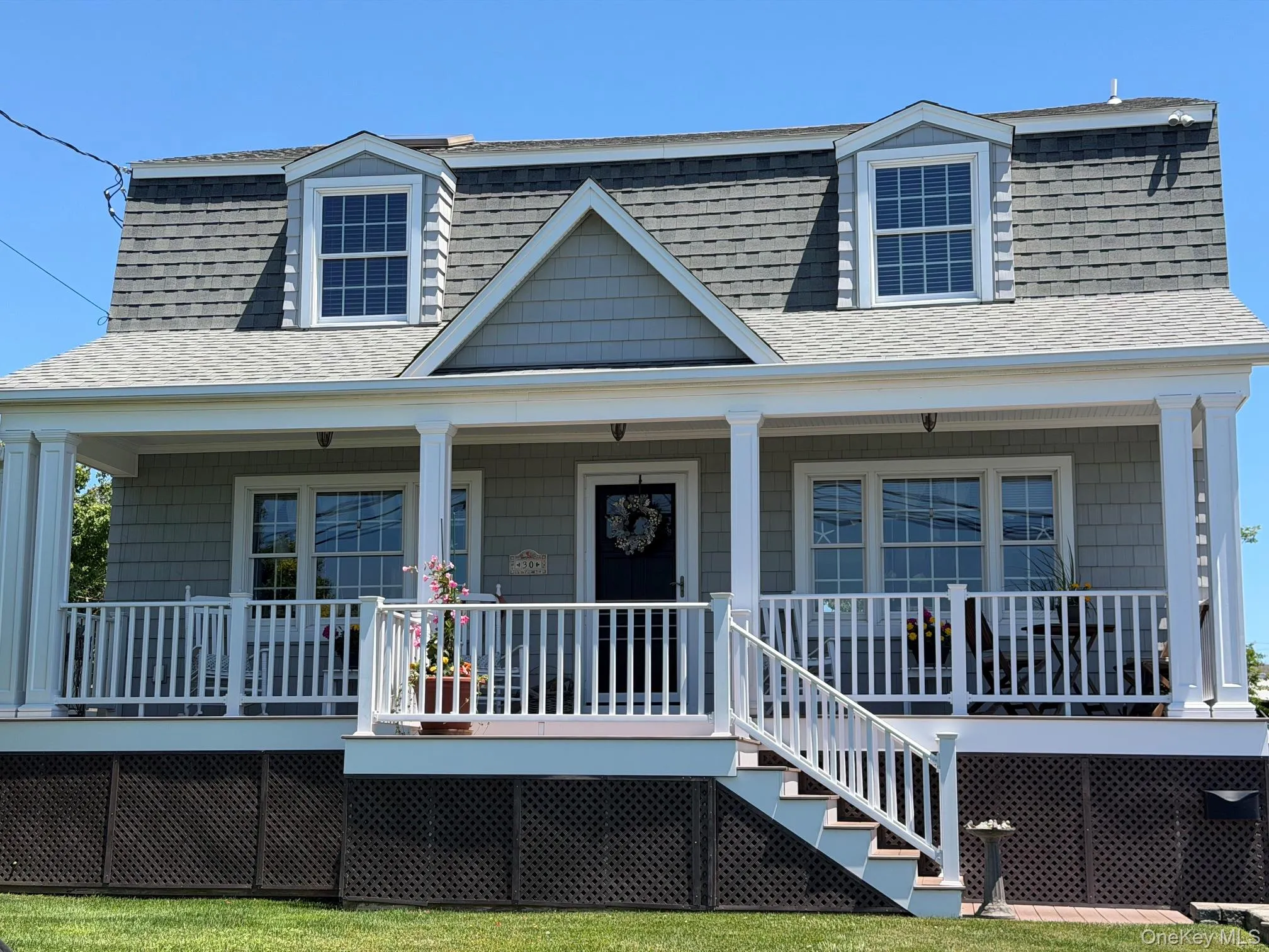 Gambrel roofline property featuring a full-width front porch with white columns, light grey shingle siding, and two dormer windows Gambrel roofline property featuring a full-width front porch with white columns, light grey shingle siding, and two dormer windows