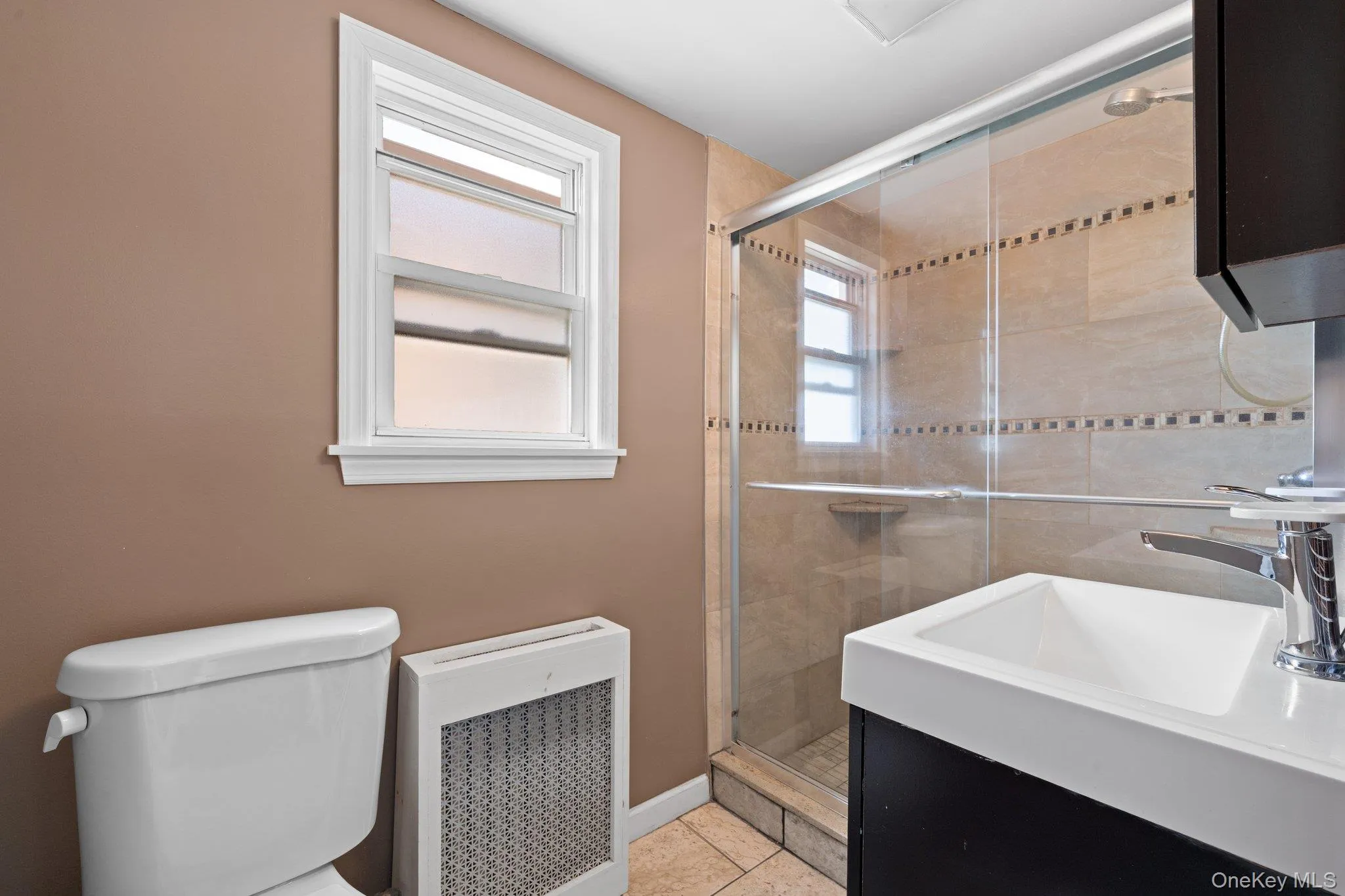 Bathroom featuring a modern shower enclosure with tiled walls, a white vanity with integrated sink, a commode, and a window with white trim Bathroom featuring a modern shower enclosure with tiled walls, a white vanity with integrated sink, a commode, and a window with white trim
