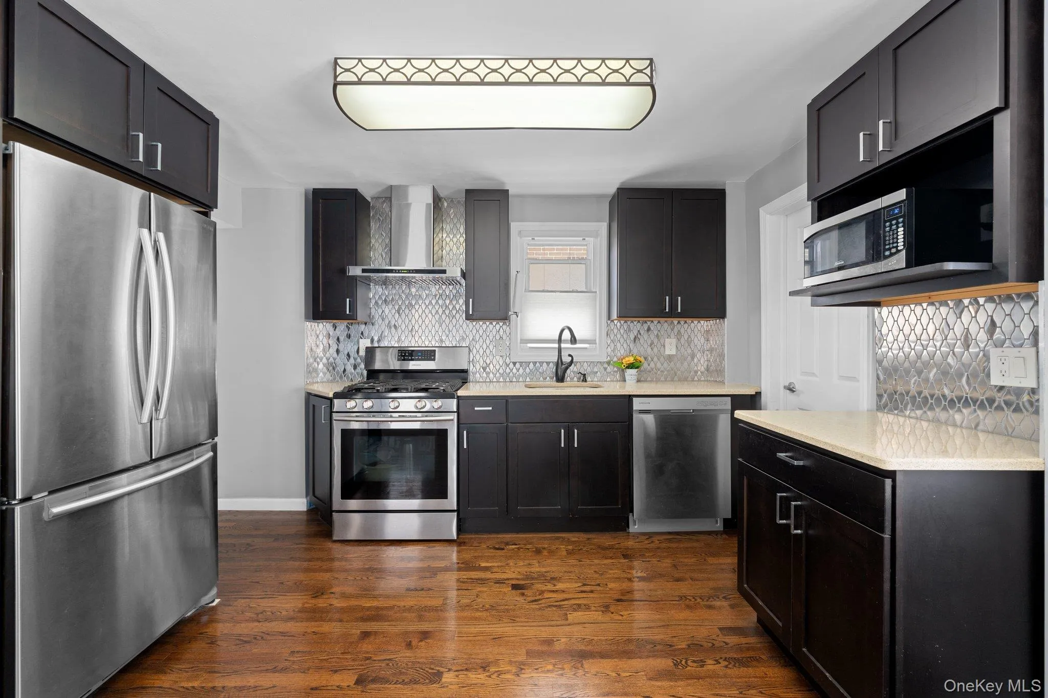 Kitchen featuring dark cabinetry, stainless steel appliances, a geometric tile backsplash, light countertops, and wood-finish flooring Kitchen featuring dark cabinetry, stainless steel appliances, a geometric tile backsplash, light countertops, and wood-finish flooring