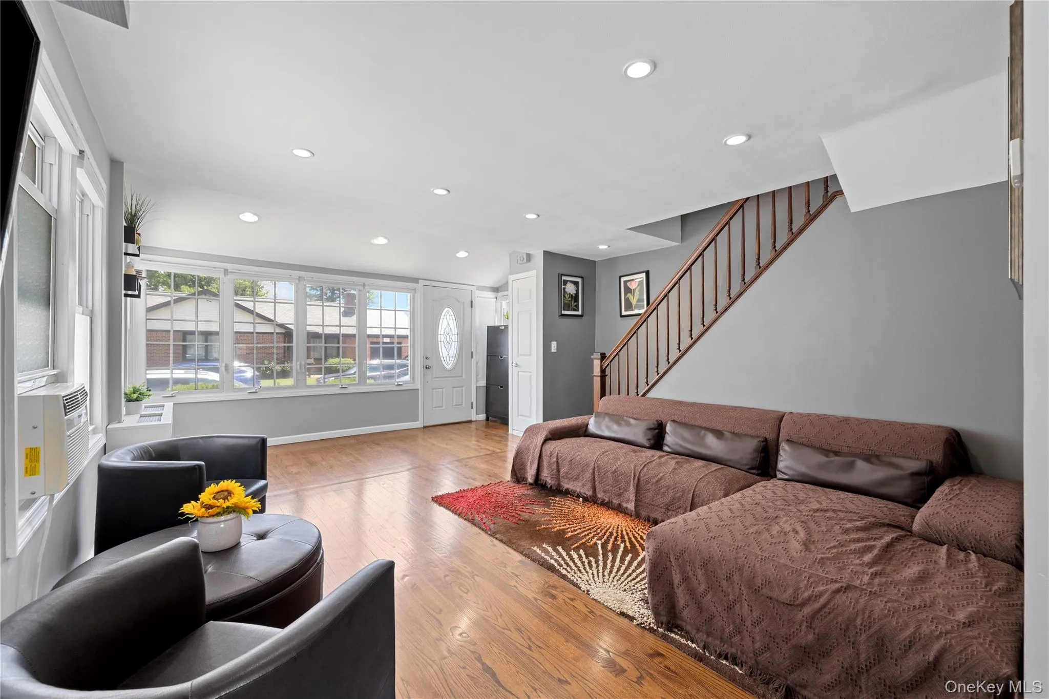 Spacious living area featuring wood-finish flooring, recessed lighting, and a prominent staircase with a wooden railing Spacious living area featuring wood-finish flooring, recessed lighting, and a prominent staircase with a wooden railing