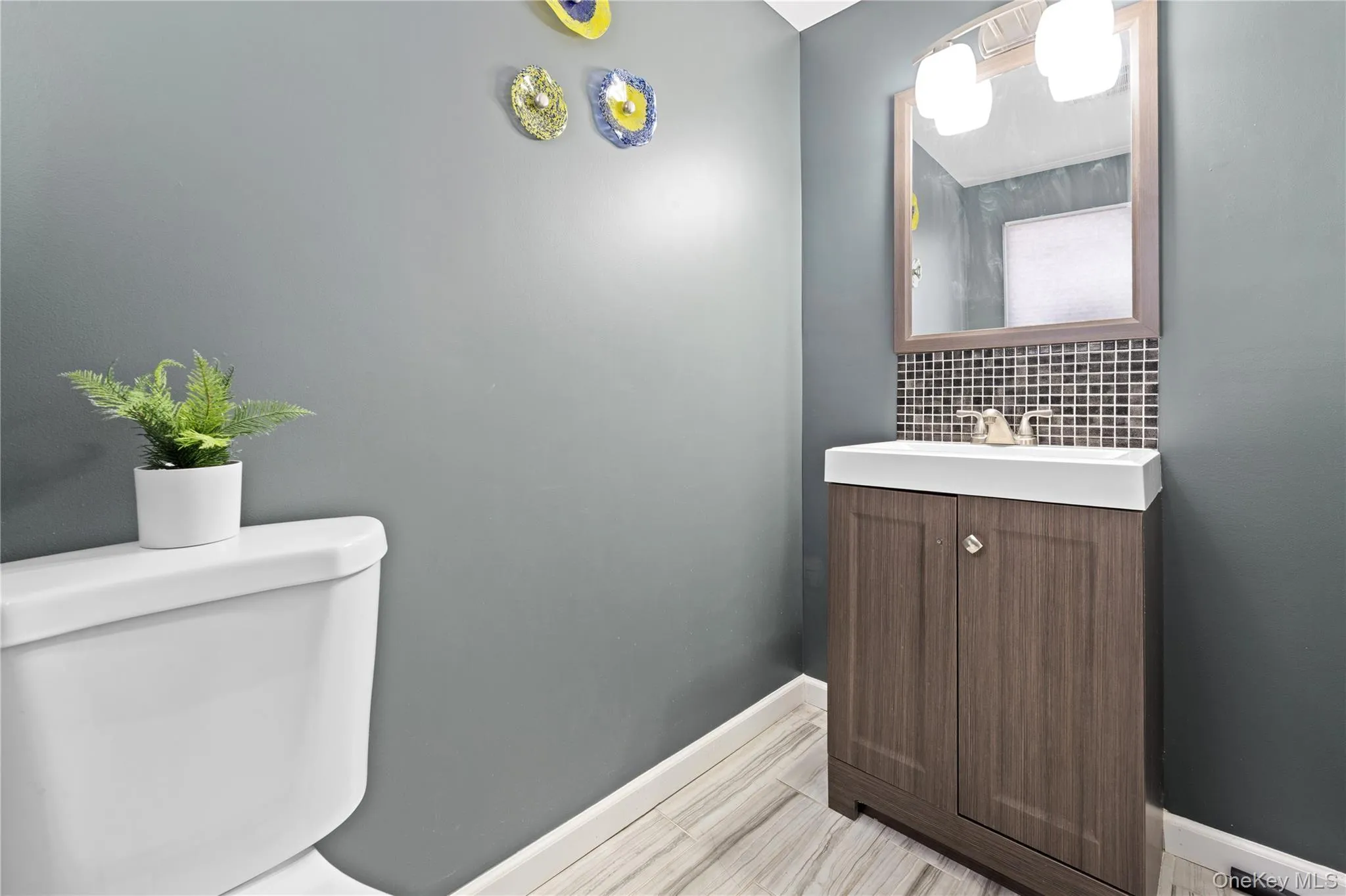 Powder room featuring a wood-finish vanity with an integrated sink, mosaic tile backsplash, and a framed mirror with overhead lighting Powder room featuring a wood-finish vanity with an integrated sink, mosaic tile backsplash, and a framed mirror with overhead lighting