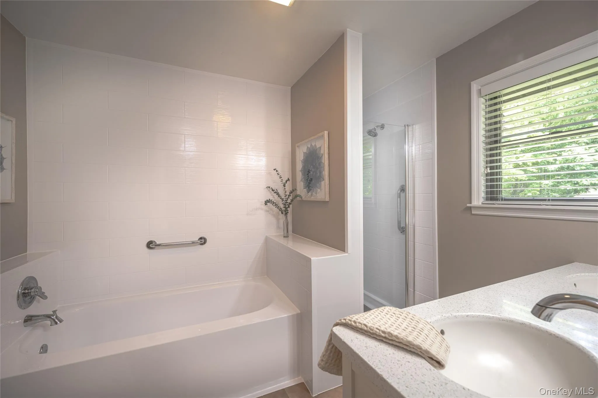 Bathroom featuring a white integrated bathtub, tiled shower enclosure, dual basin vanity with a light countertop, and a window with blinds Bathroom featuring a white integrated bathtub, tiled shower enclosure, dual basin vanity with a light countertop, and a window with blinds
