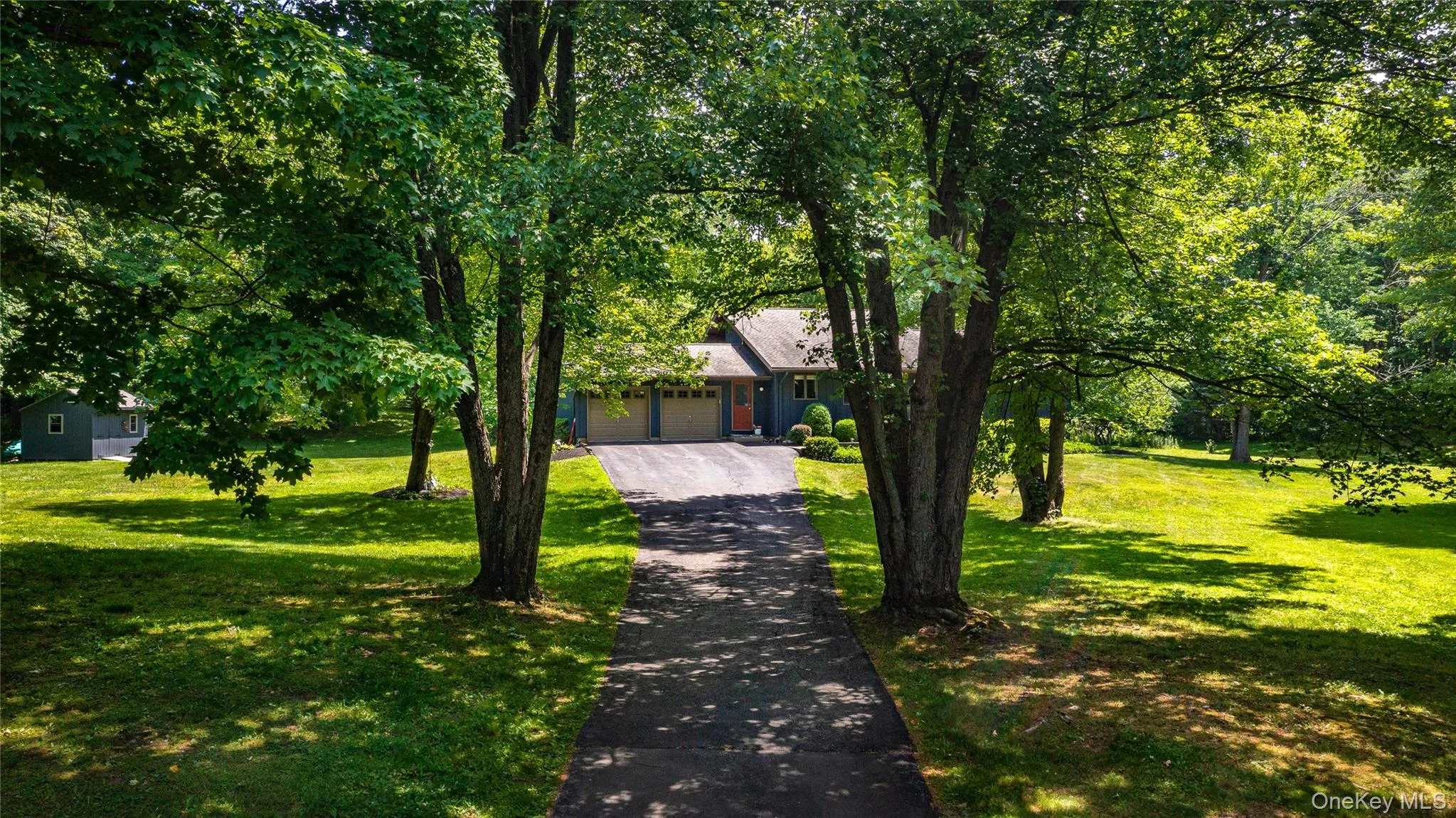 Spacious grounds featuring a paved driveway framed by mature trees, a lush green lawn, and a multi-car garage Spacious grounds featuring a paved driveway framed by mature trees, a lush green lawn, and a multi-car garage
