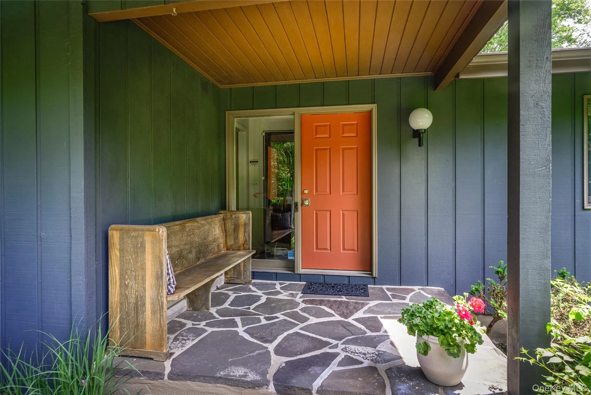 Inviting front entryway featuring a vibrant orange door, natural wood plank ceiling, flagstone-style porch, and vertical exterior siding Inviting front entryway featuring a vibrant orange door, natural wood plank ceiling, flagstone-style porch, and vertical exterior siding