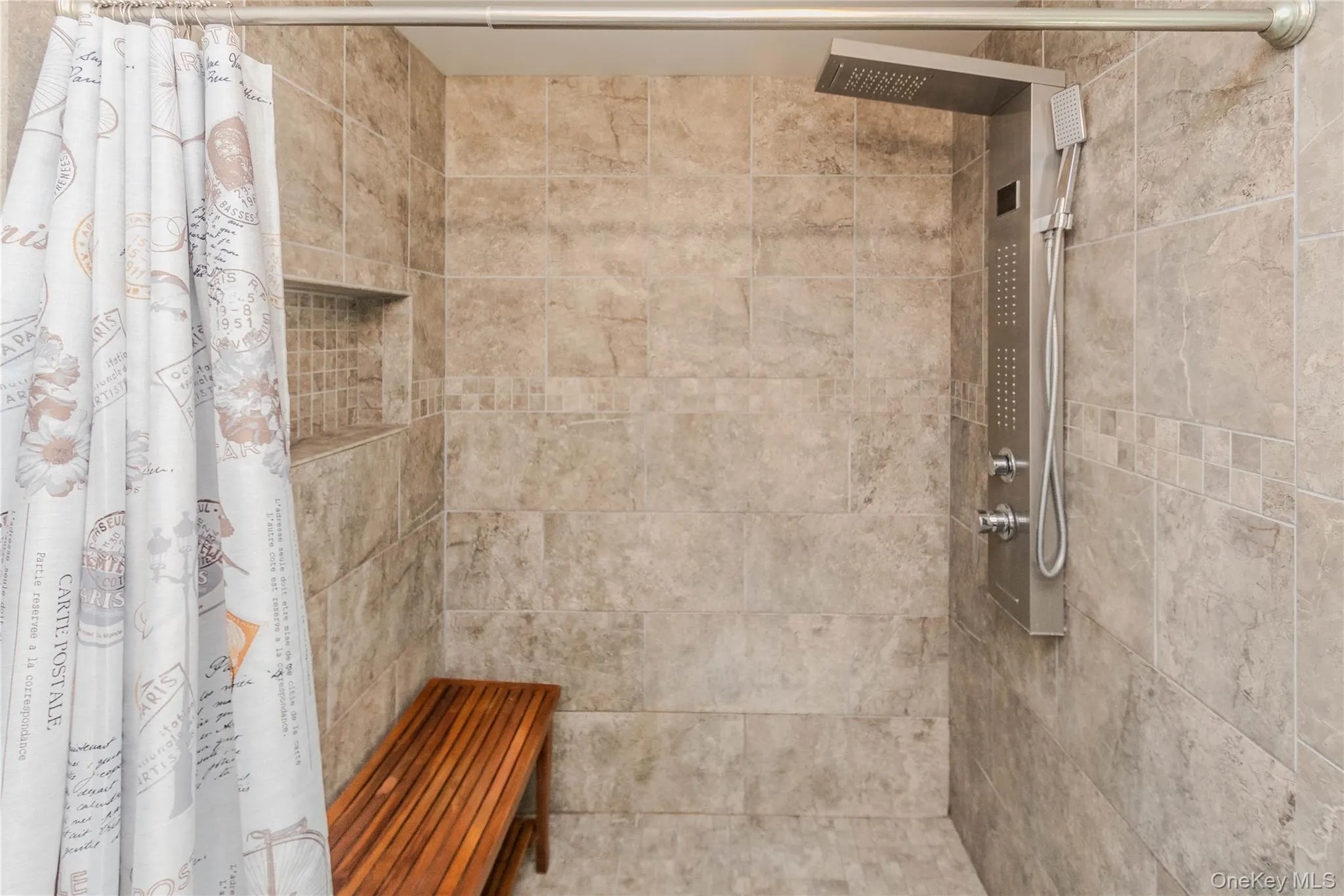 Shower stall featuring large format tile walls, a built-in niche with mosaic tile accents, a multi-function shower panel with an overhead rain shower and handheld sprayer, and a slatted wood bench Shower stall featuring large format tile walls, a built-in niche with mosaic tile accents, a multi-function shower panel with an overhead rain shower and handheld sprayer, and a slatted wood bench